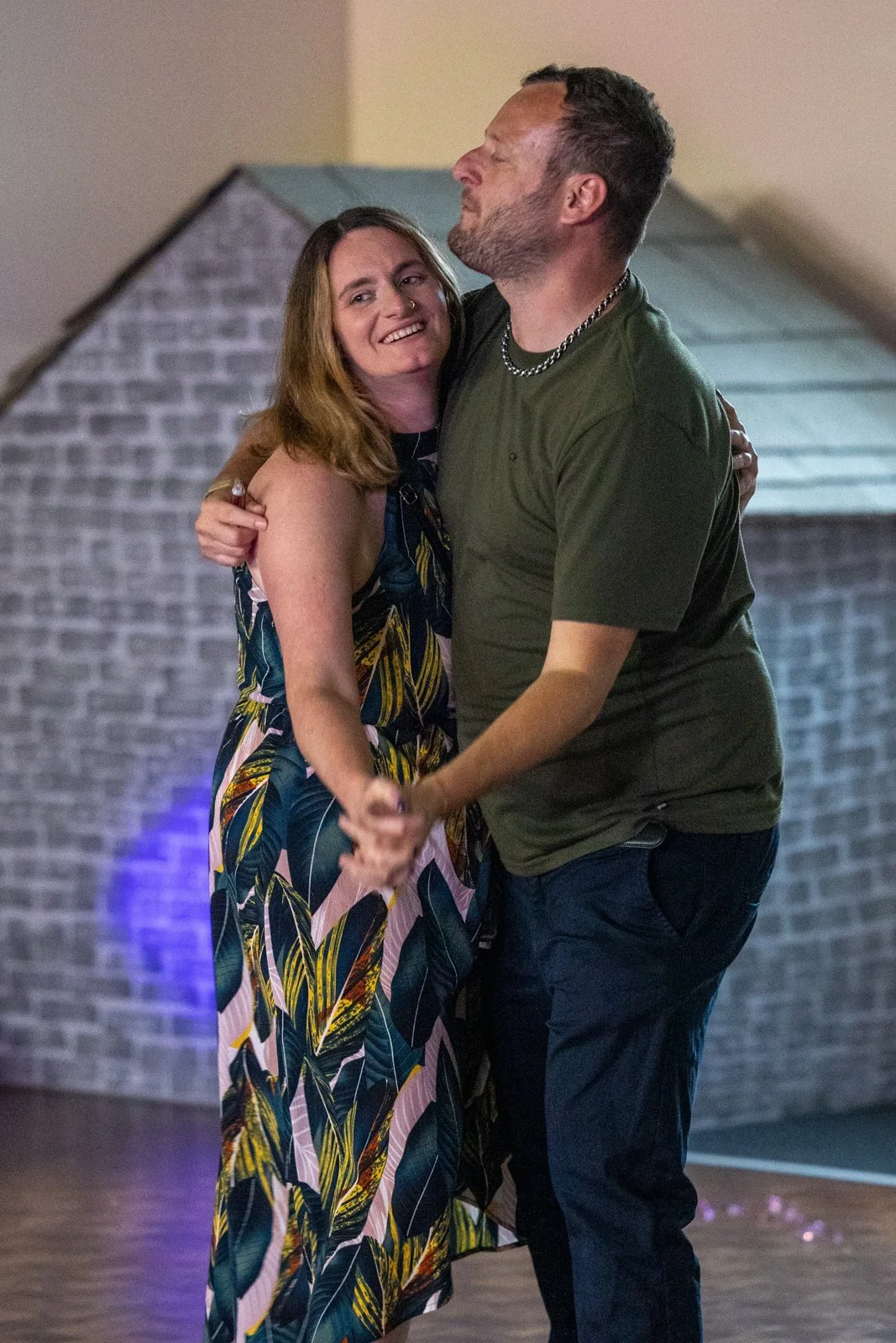 A man and woman are dancing together, smiling and embracing in a warmly lit room with a brick wall in the background.