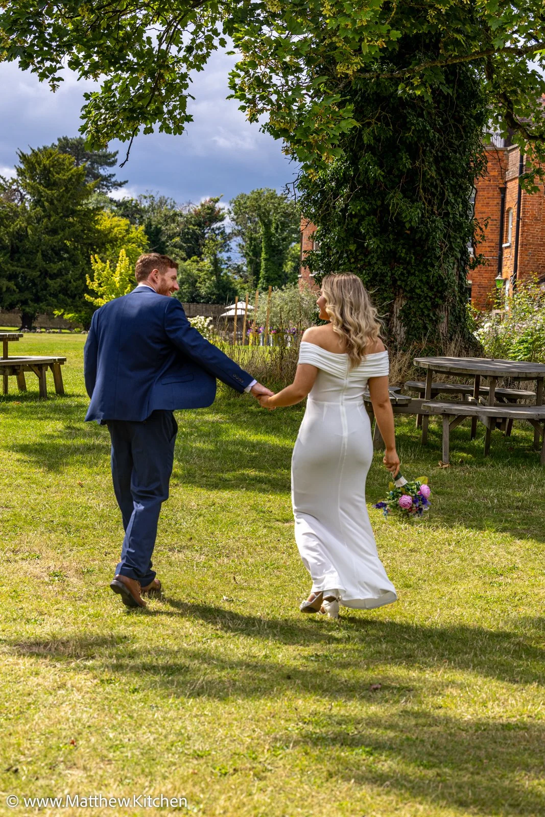 A bride and groom holding hands and walking on a grassy area outdoors during daytime. The bride wears a white off-the-shoulder wedding dress and holds a bouquet of flowers. The groom is dressed in a blue suit. There are picnic tables and a large tree