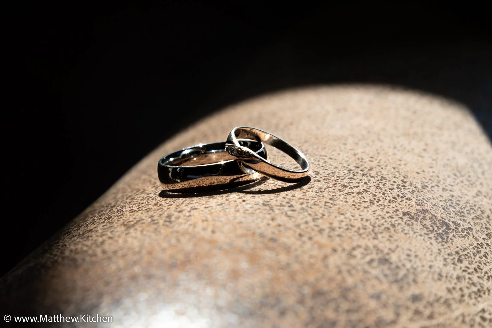 Two wedding bands resting on a textured surface illuminated by sunlight with dark shadow background.