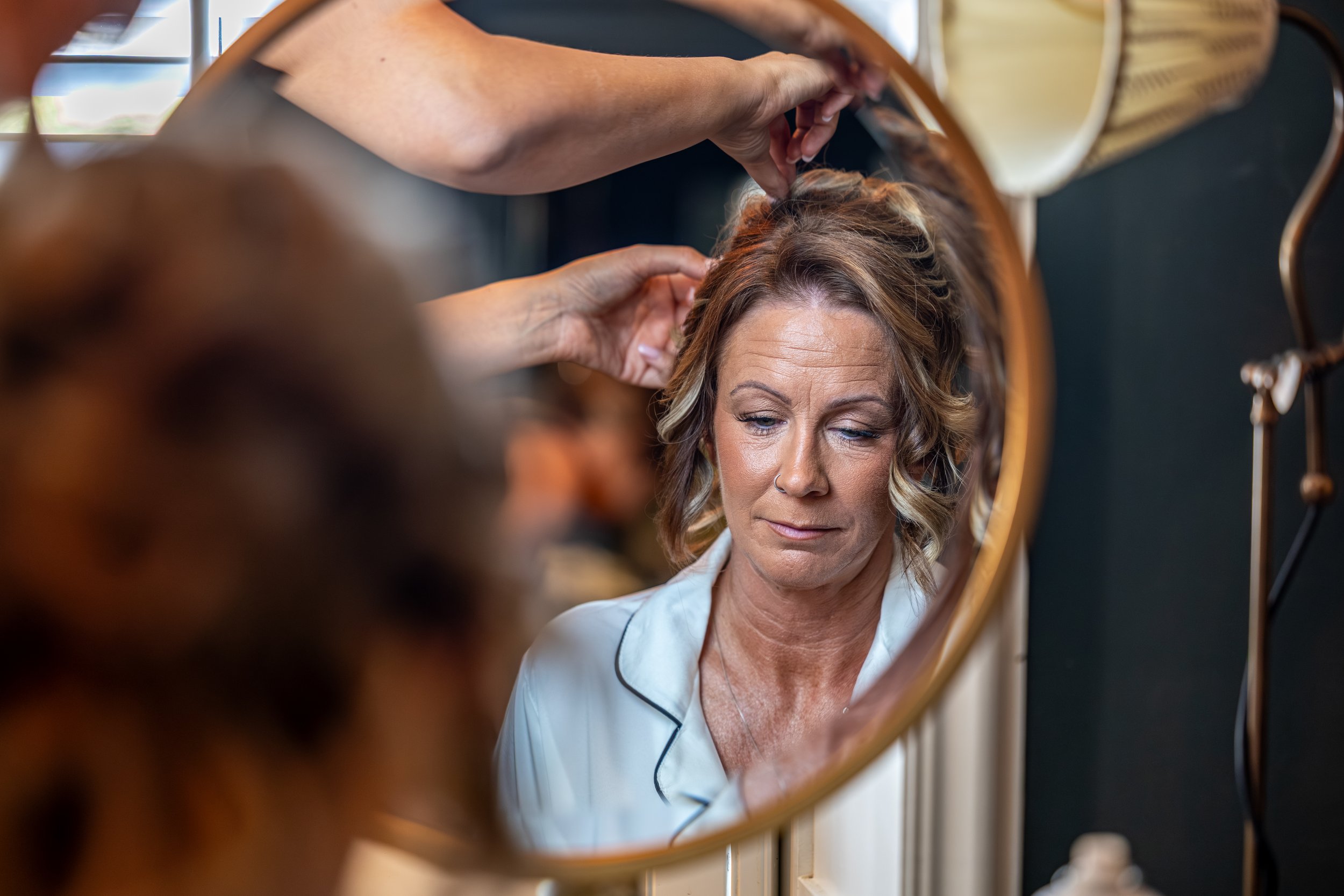 A Bride with wavy blonde hair and light skin, wearing a white shirt, looking into a round mirror while a stylist styles her hair. The scene is captured through the mirror, showing the woman's face and the stylist's hands.