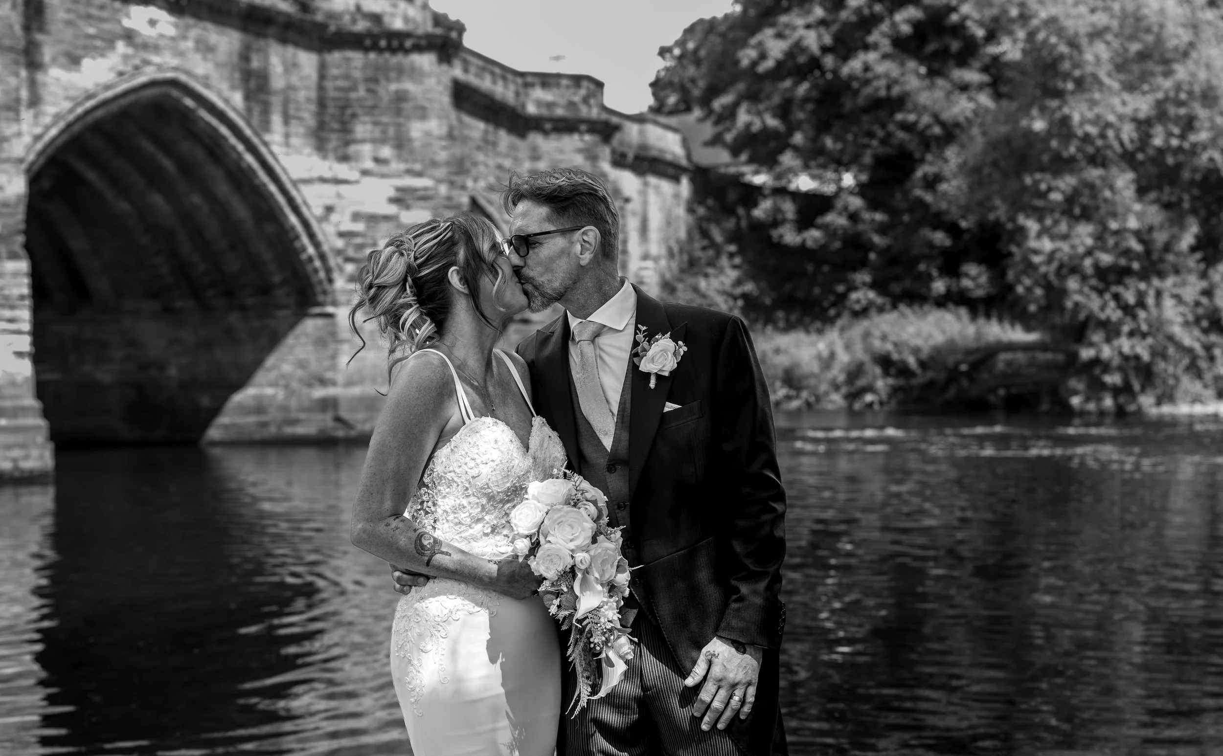 A black-and-white photo of a bride and groom kissing outdoors by a river with a stone bridge and trees in the background.