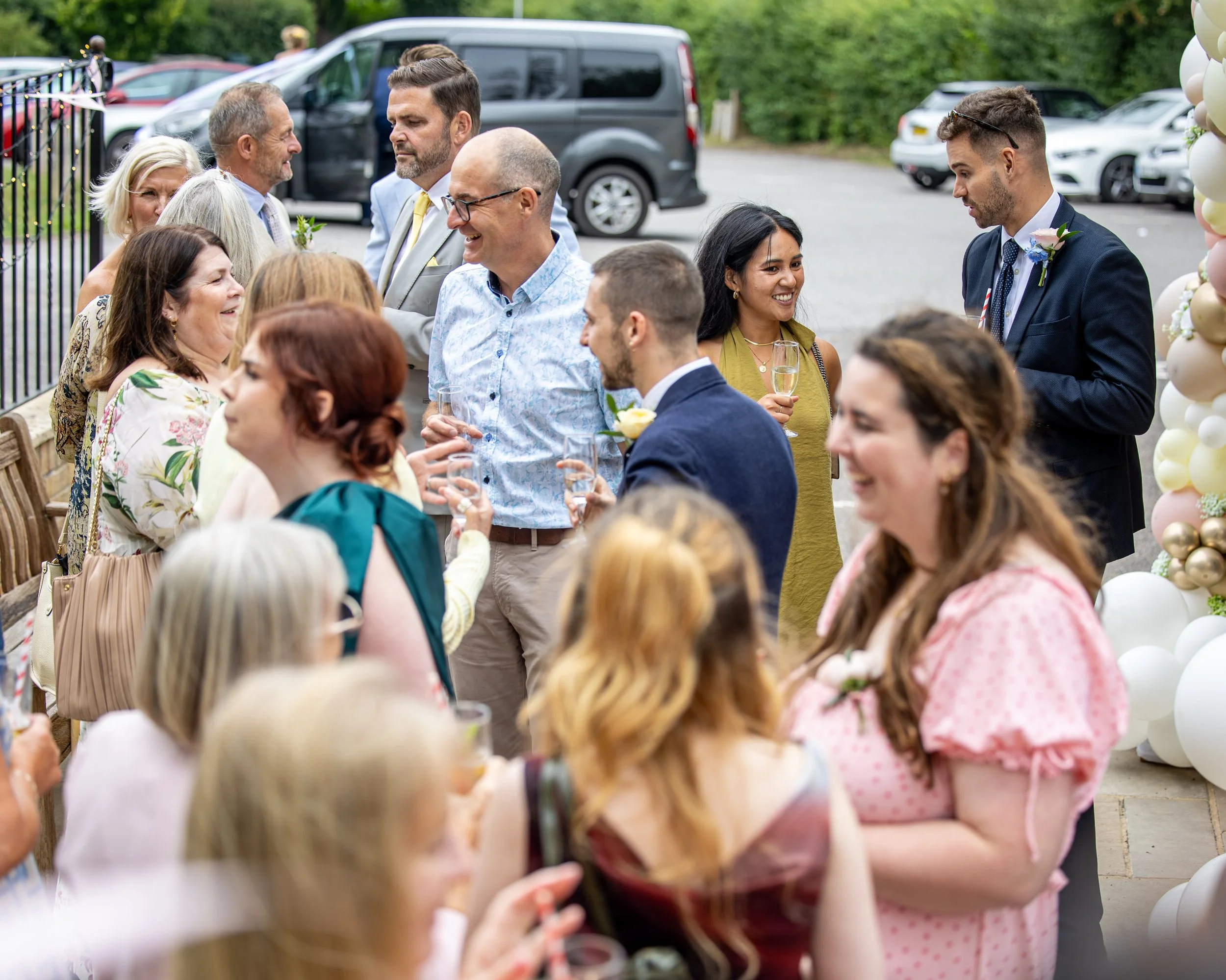 People gathered outdoors for a celebration, talking and holding glasses of champagne, with some wearing formal attire and mingling near a decorated balloon arch.