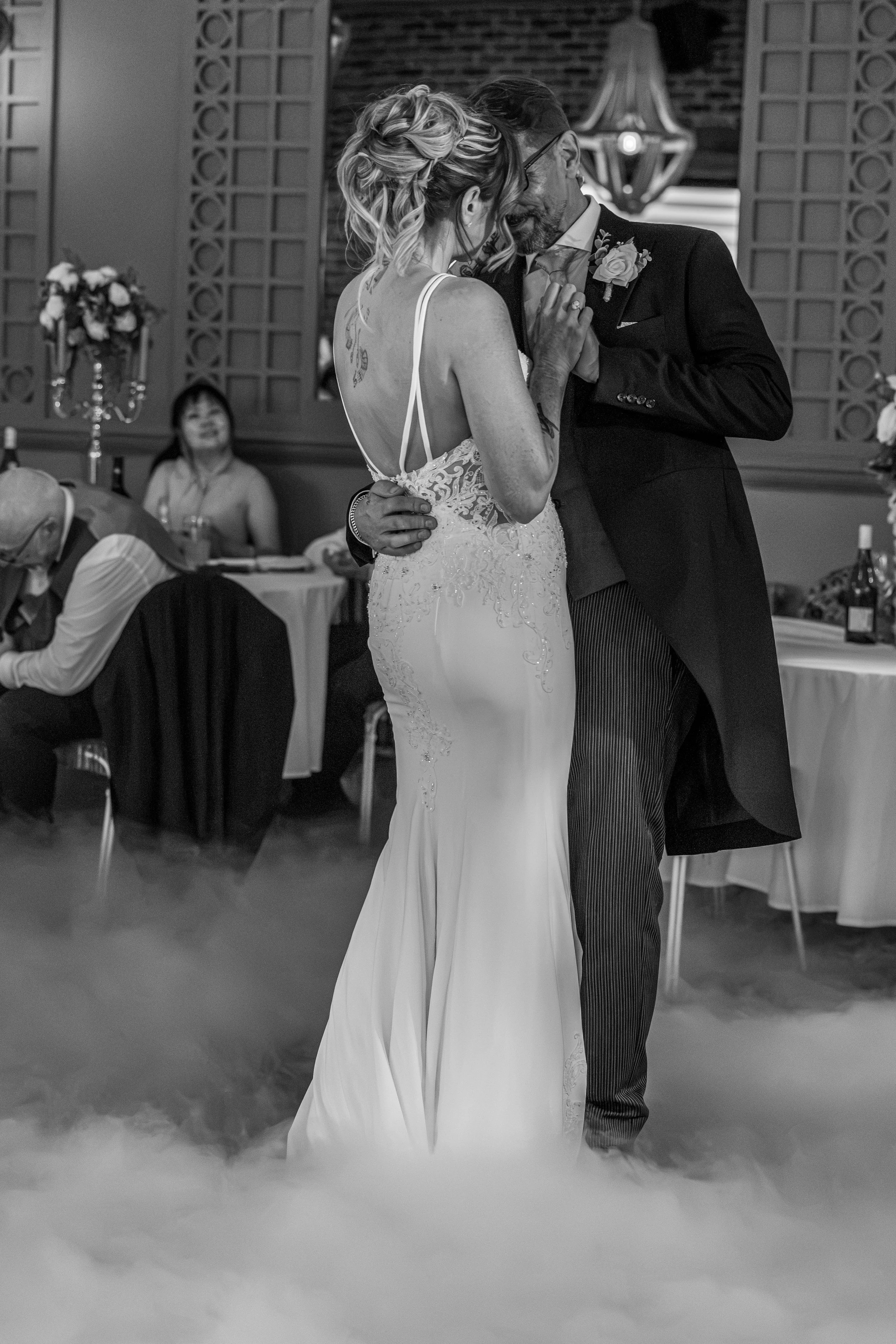 A bride and groom dancing at their wedding reception, with guests seated at tables in the background.