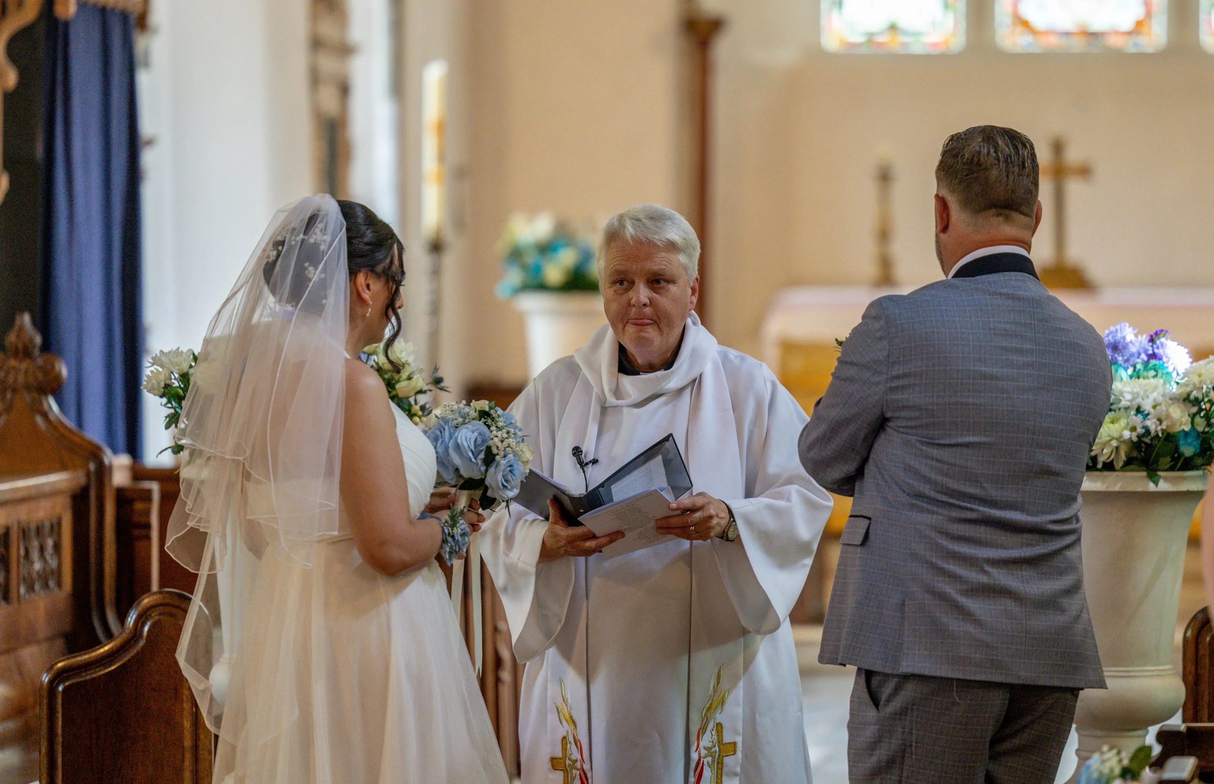 A woman in a white wedding dress and veil, a man in a gray suit, and a priest are participating in a wedding ceremony inside a church, with stained glass windows in the background.