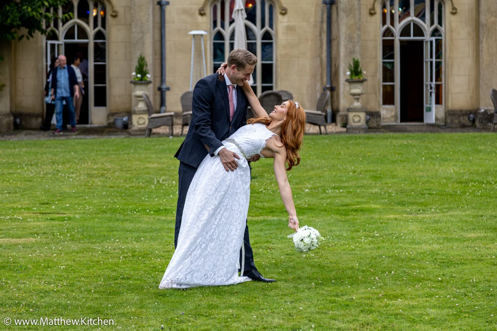 A newlywed couple dancing outside on a green lawn, with the groom in a dark suit and the bride in a white lace wedding dress, holding a bouquet of white flowers. In the background, a building with large arched windows and a few people near the door.