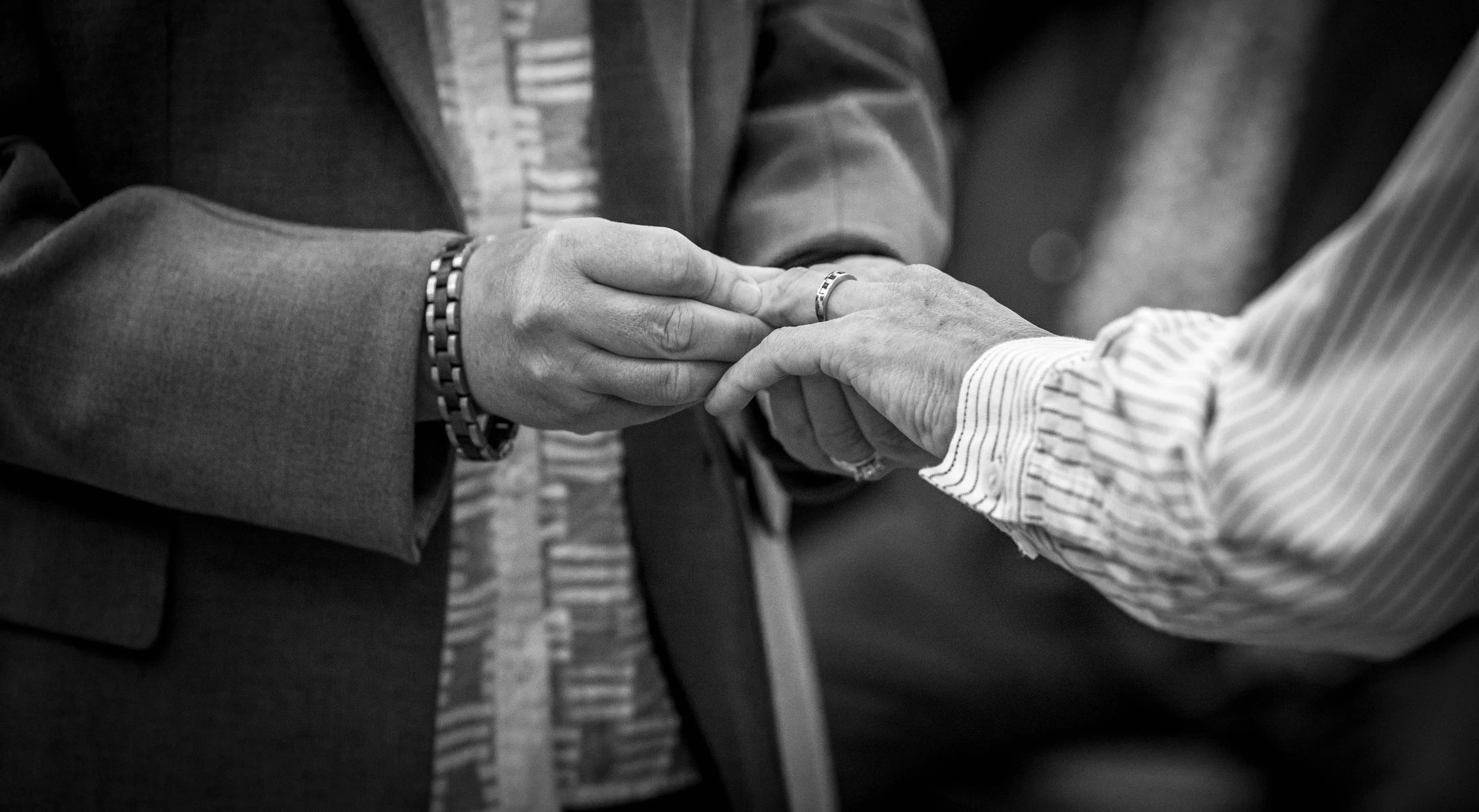 Two people holding hands during a ceremony, with one person wearing a suit and a wristwatch, the other wearing a striped shirt, symbolic of a wedding or commitment event.
