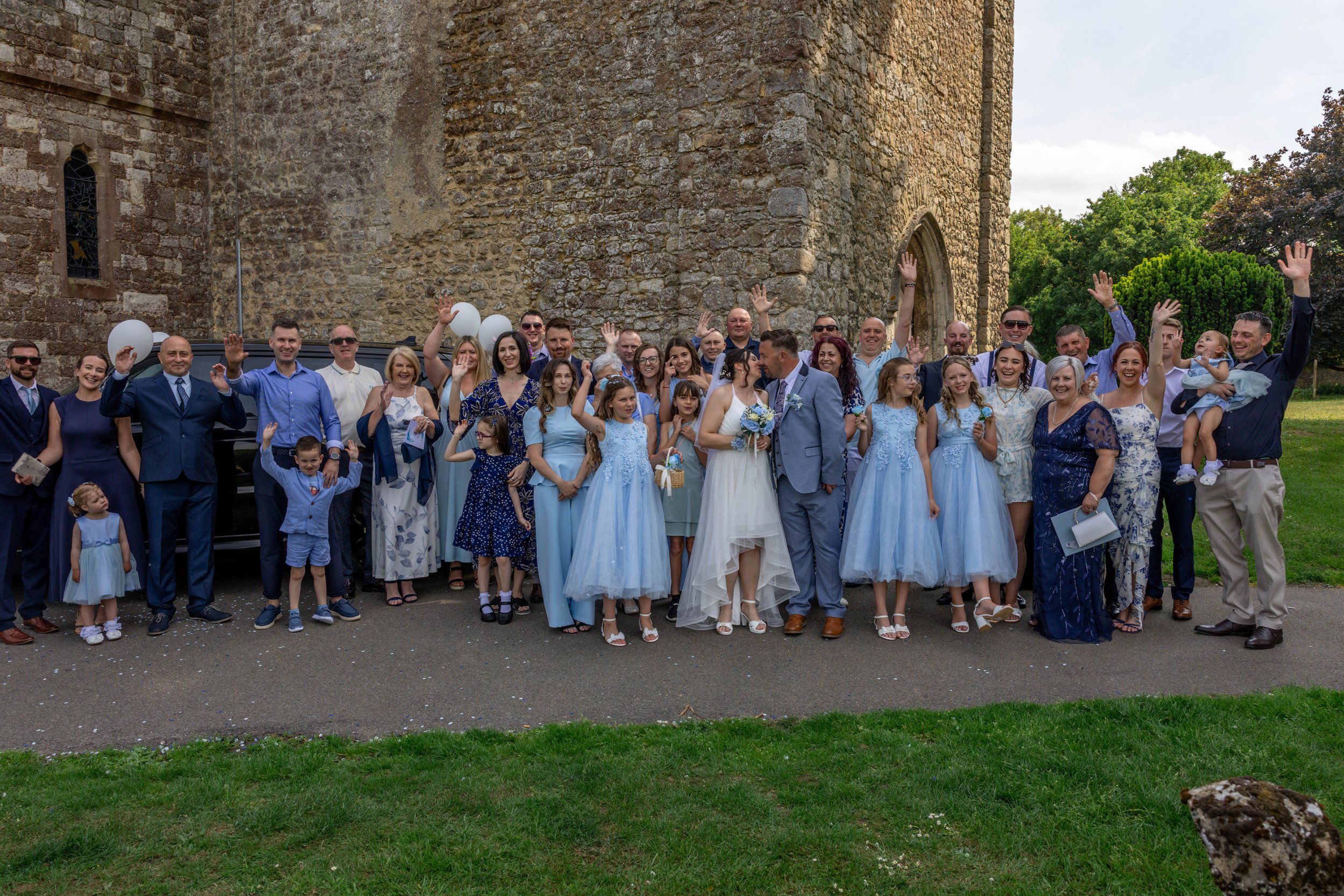 A large group of people, including children and adults, dressed in formal attire, gathering outside near a stone church for a celebration or wedding, with some waving and smiling.