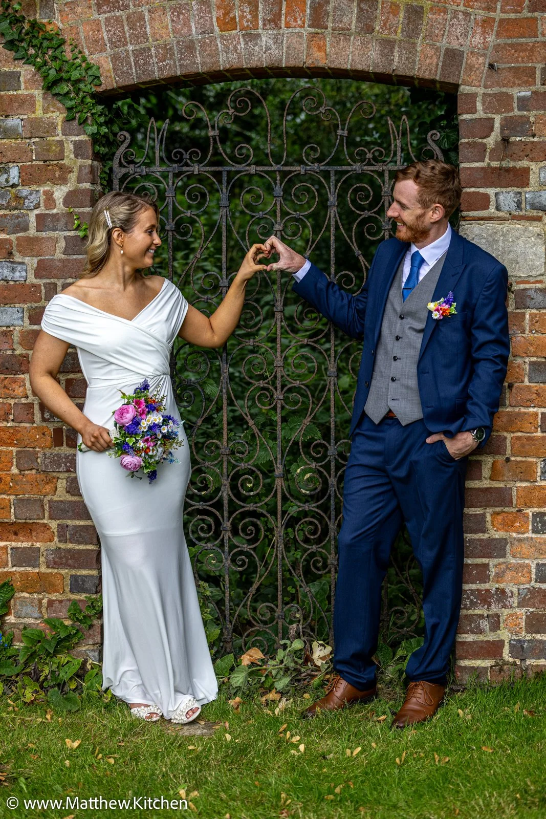 A bride and groom dressed in wedding attire stand next to a brick wall and iron gate, holding hands and smiling.