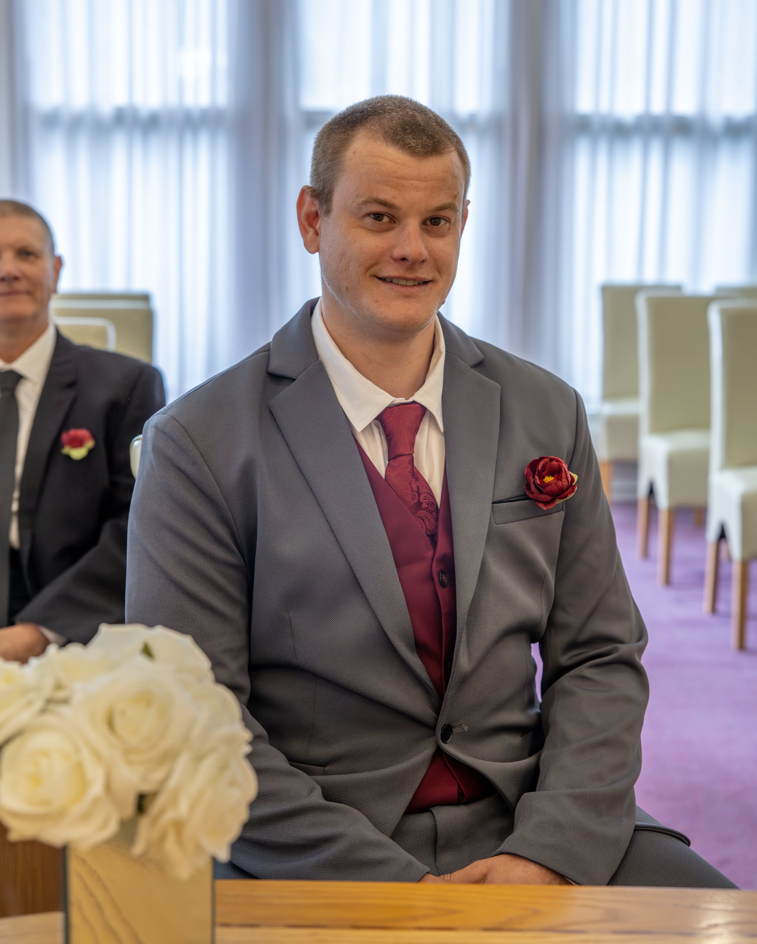 A groom in a gray suit and red vest smiling and sitting at a table with a blurred background of chairs and windows, with a bouquet of white roses in the foreground.