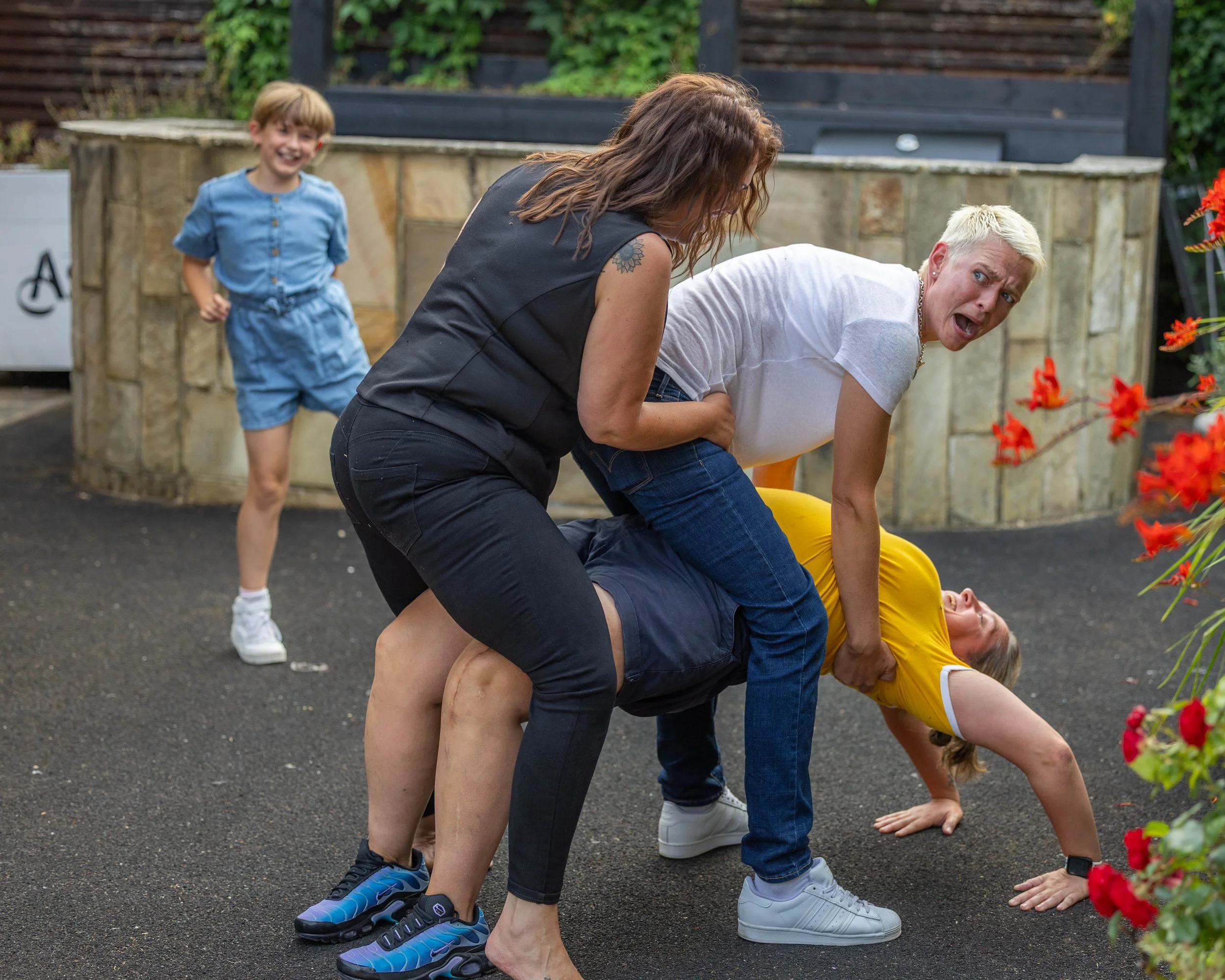 Children playing a game of human lawn chairs, with one woman leaning back and others supporting her, while two children observe and laugh in the background.