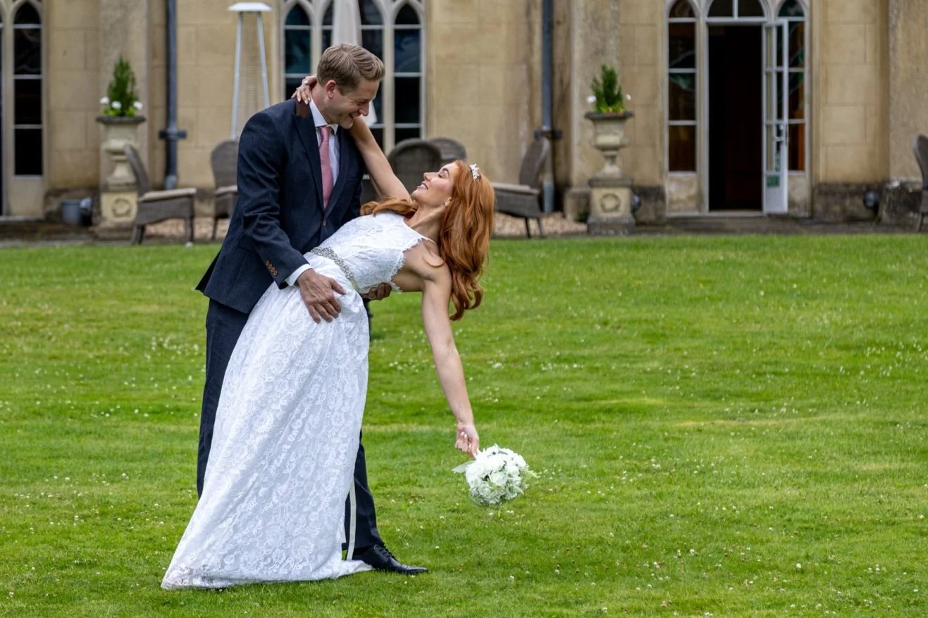 A couple, dressed in wedding attire, dancing on a grassy field outside a large stone building, with the groom dipping the bride while she holds a bouquet of flowers.