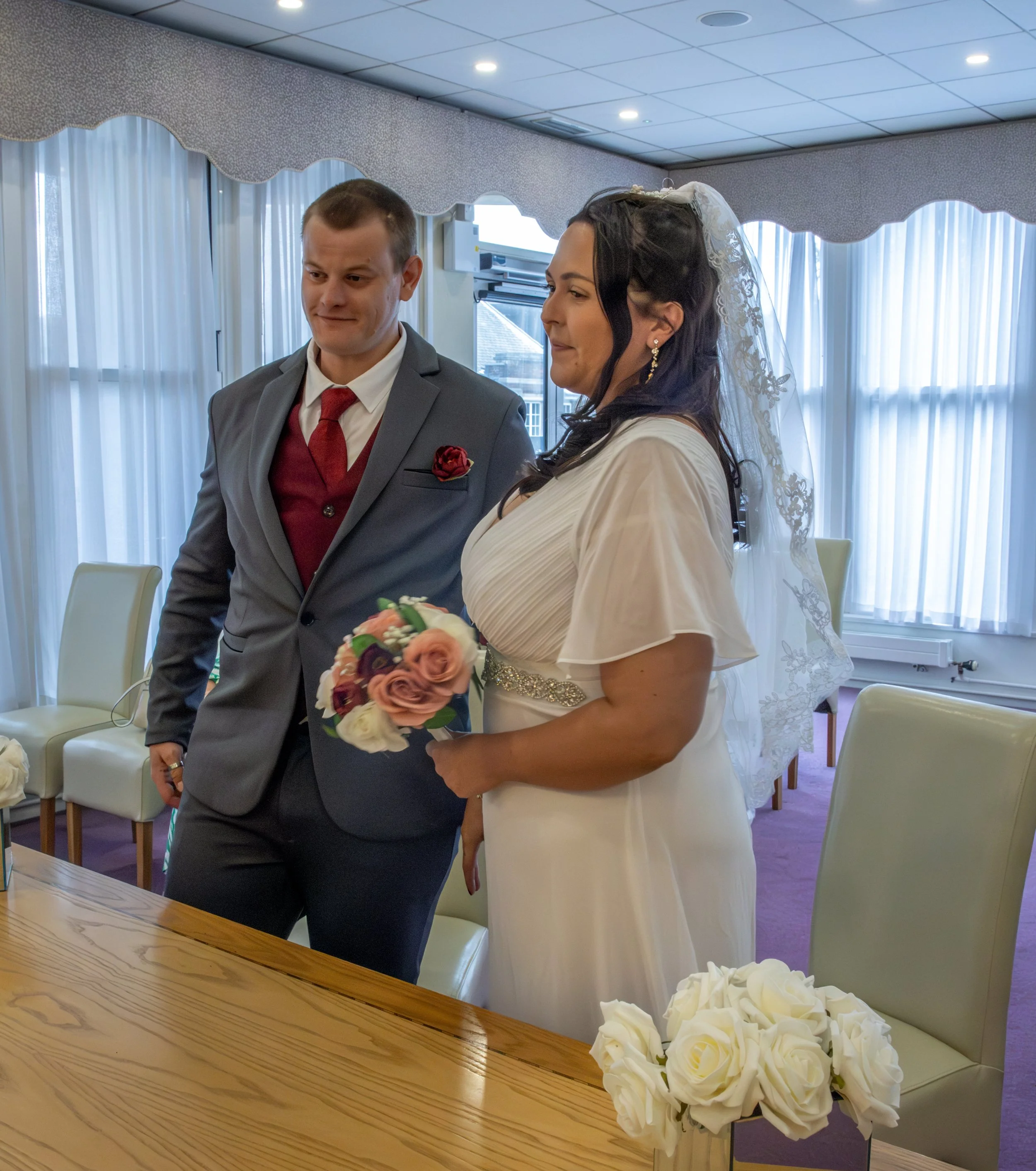 A bride and groom during a wedding ceremony indoors. The groom is wearing a gray suit with a red tie and a red rose boutonniere. The bride is wearing a white dress with lace details and a veil with lace embroidery. She is holding a bouquet of pink an