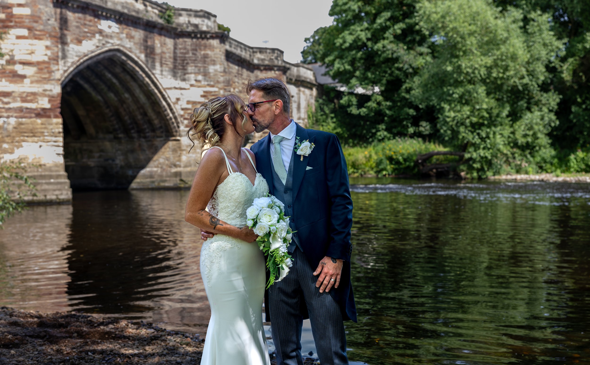 A bride and groom kiss by a river with a stone bridge and green trees in the background.