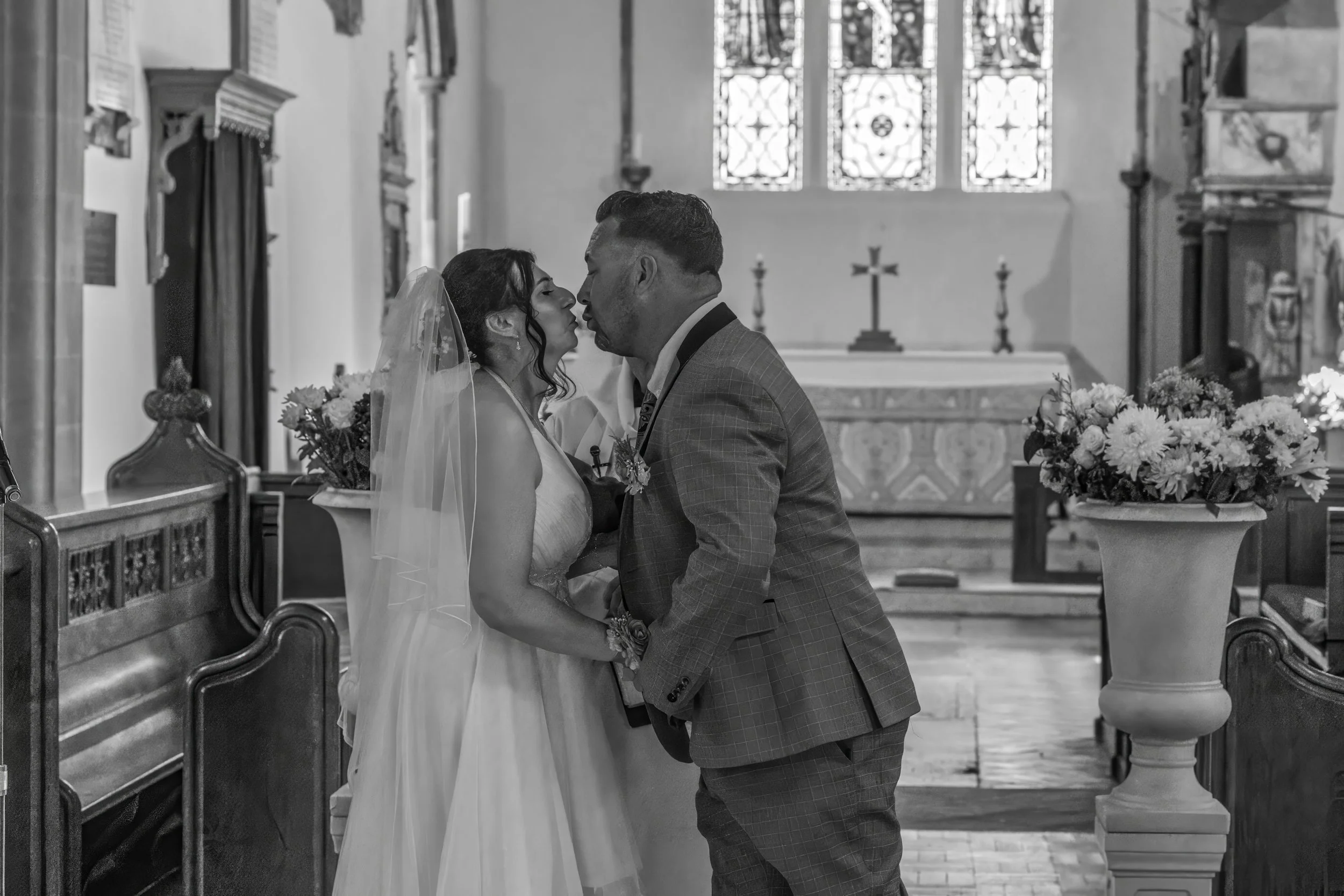 Black and white photo of a bride and groom kissing inside a church, with stained glass windows and floral arrangements.