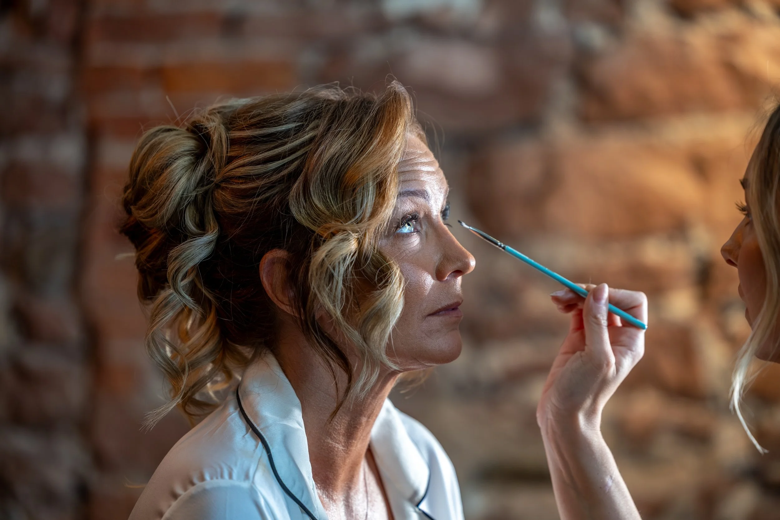 A Bride with curly blonde hair getting her makeup done by a makeup artist.