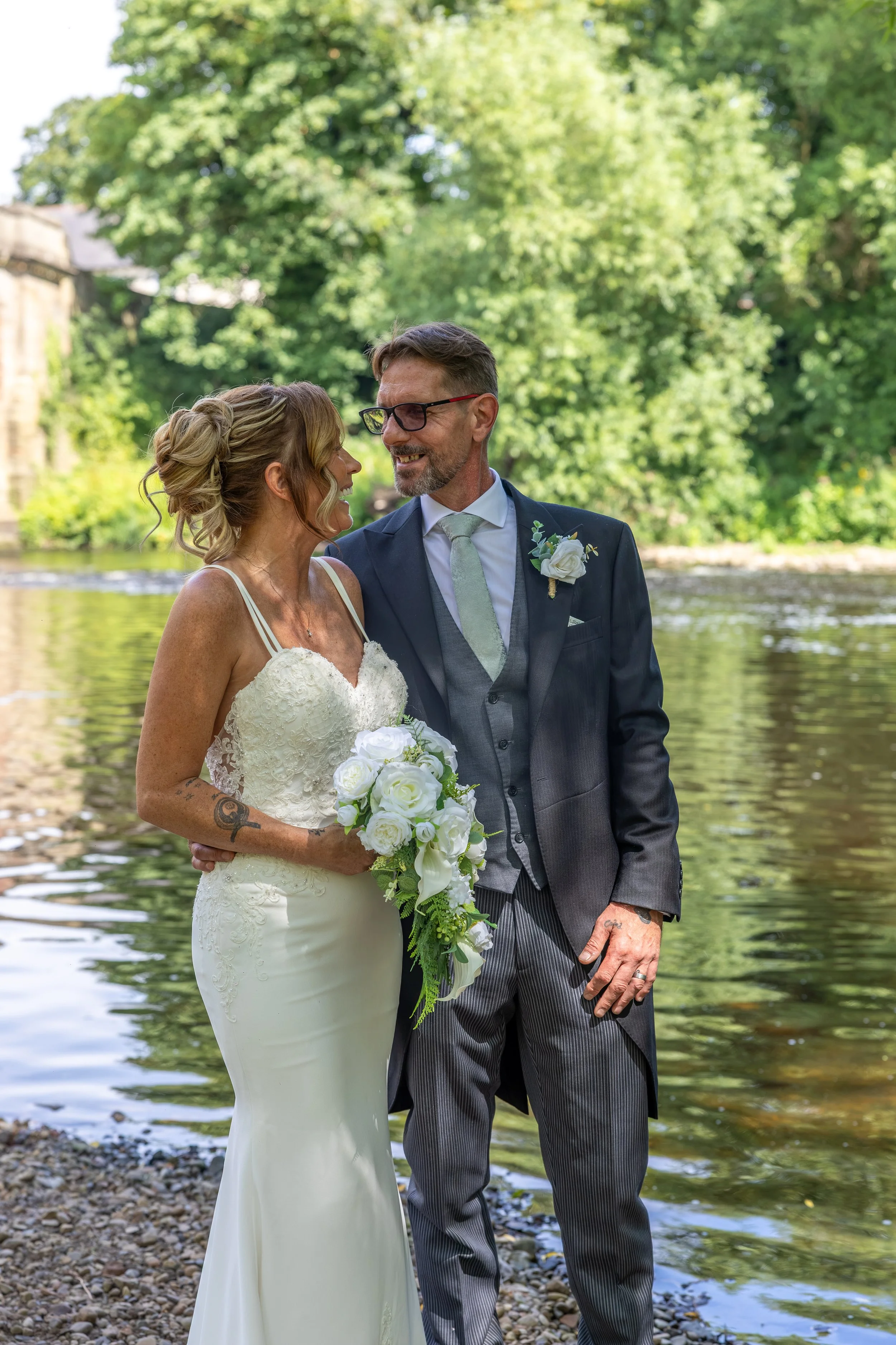 A bride and groom stand close together outdoors near a river, smiling at each other. The bride wears a white lace wedding gown and holds a bouquet of white roses, while the groom wears a dark suit with a white shirt, a light-colored tie, and a bouton