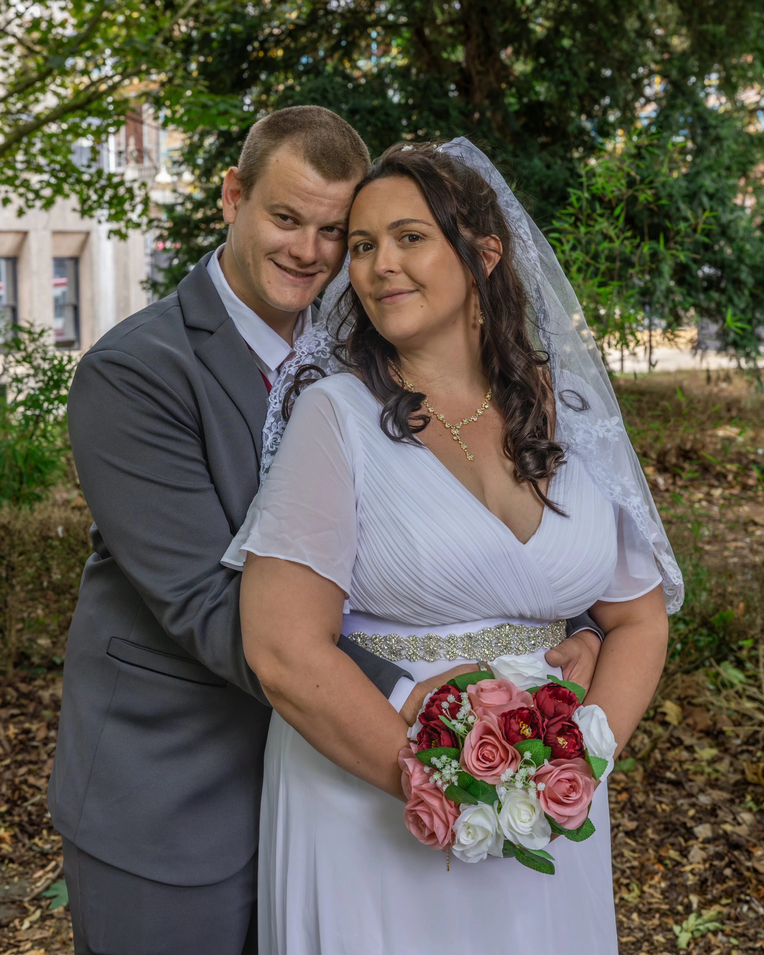 A newlywed couple stands outdoors, with the groom in a gray suit and the bride in a white dress holding a bouquet of pink, red, and white flowers, surrounded by green trees and a partly cloudy sky.