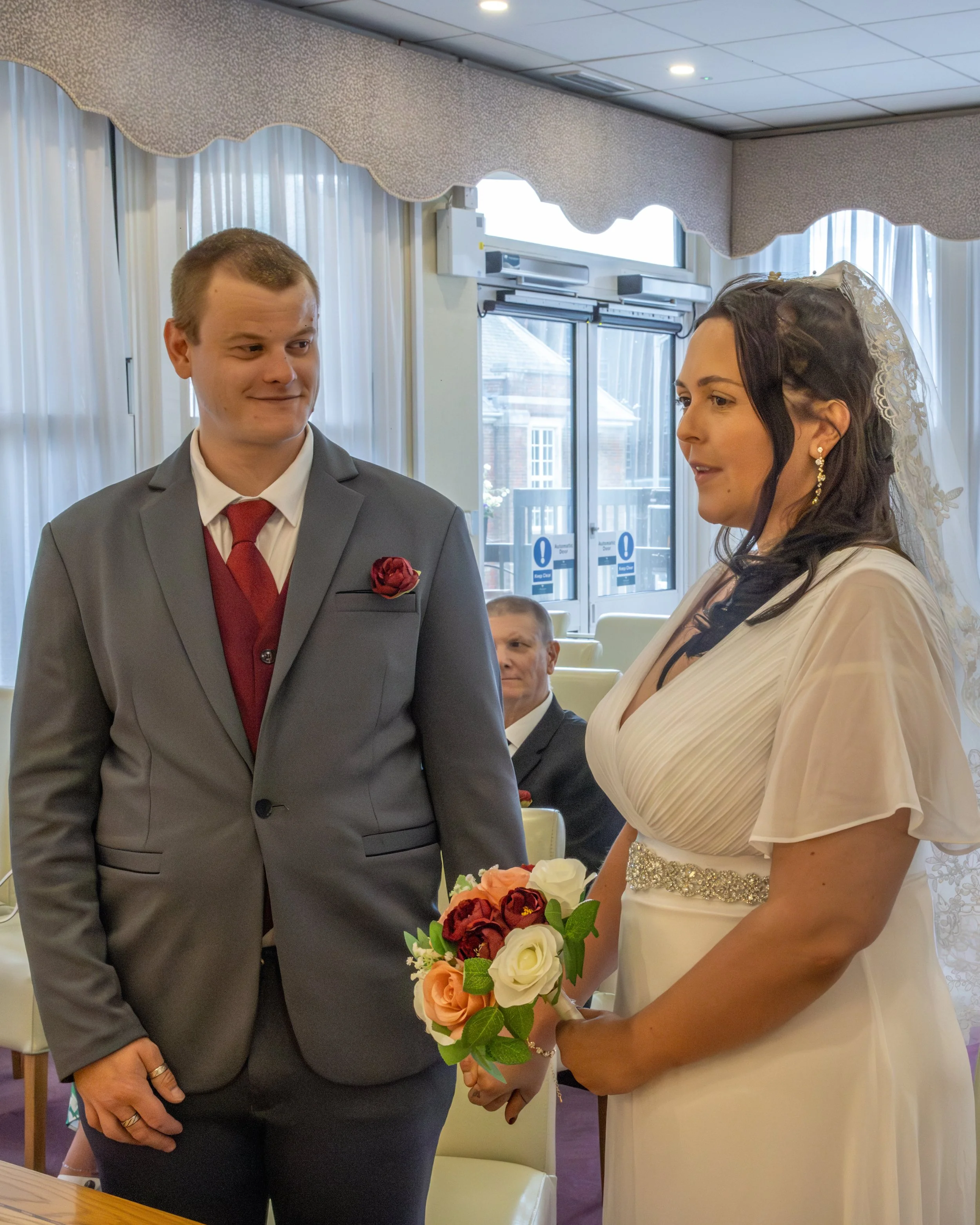 A bride with dark hair wearing a white dress and veil holding a bouquet of roses, standing next to a groom with short light hair wearing a gray suit and red tie, at a wedding ceremony.