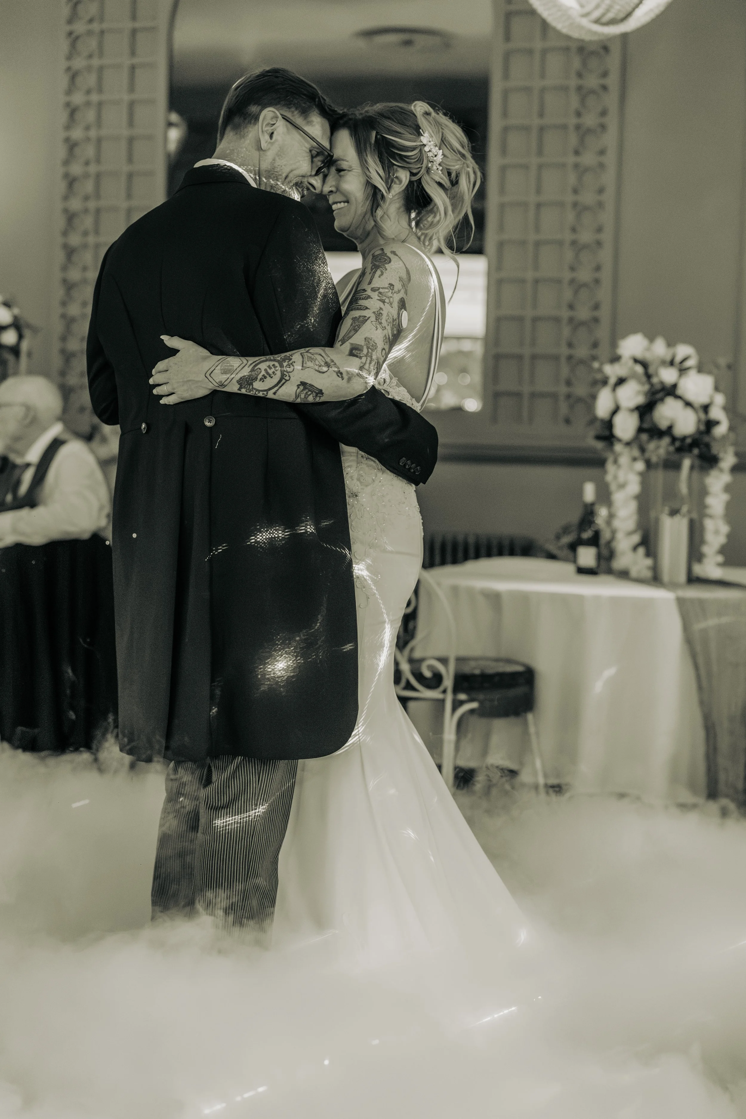 Black and white photo of a wedding reception, showing a bride and groom hugging and smiling. The groom is wearing glasses and a dark suit, the bride has tattoos and is wearing a wedding dress. They are dancing and look happy.
