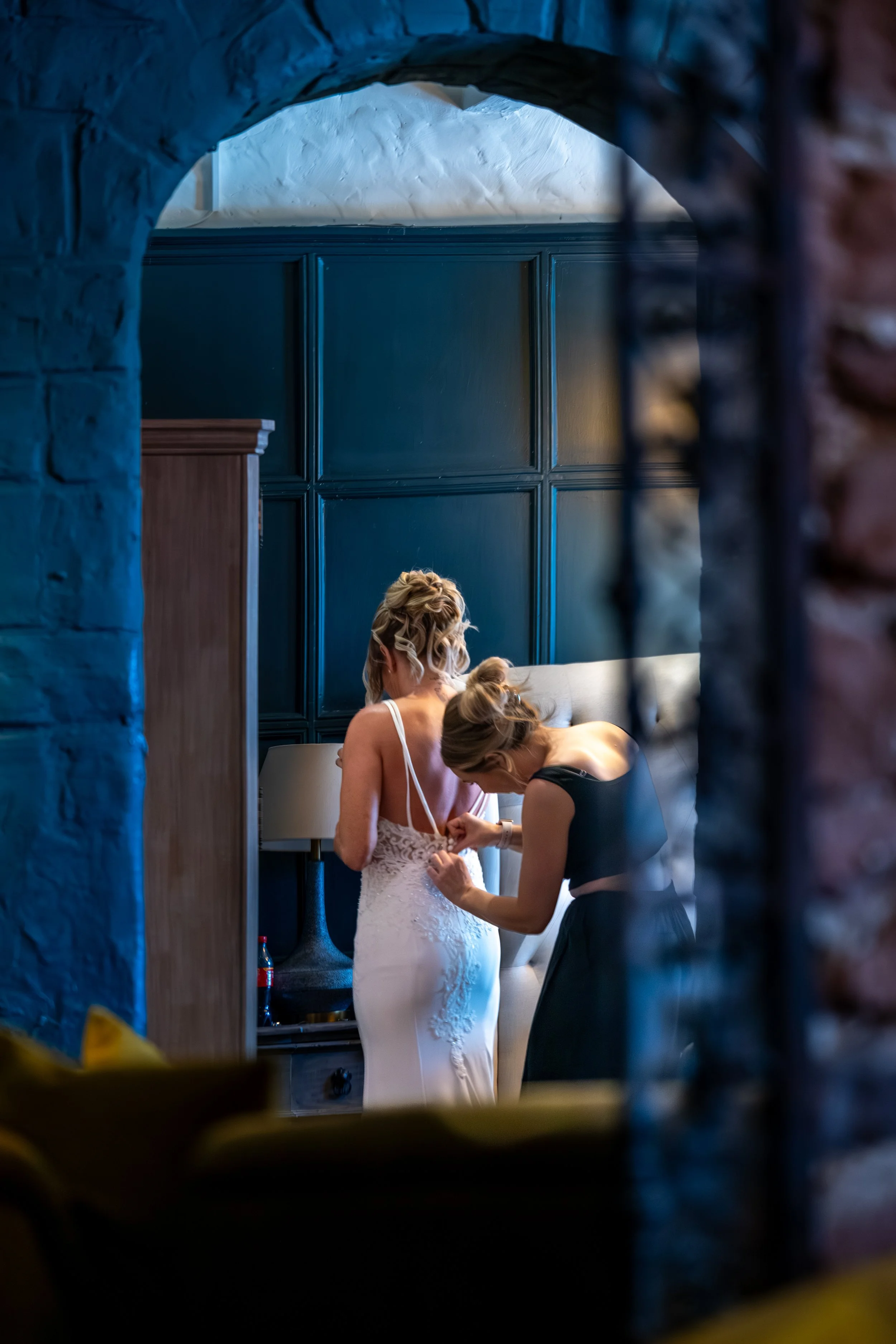 A bride in a white wedding dress is getting ready, assisted by another woman, in a dimly lit room with blue-paneled walls.