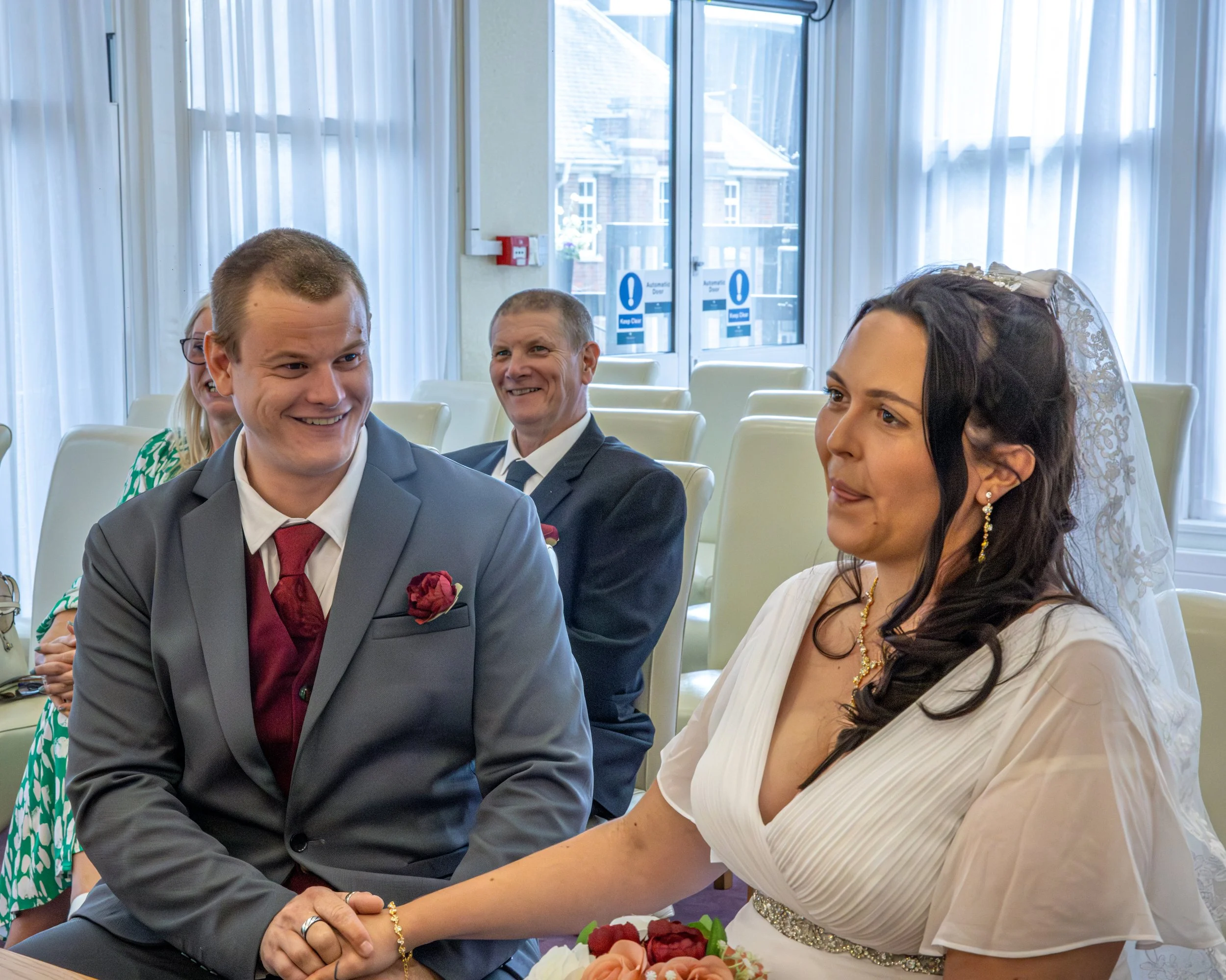A wedding ceremony with a groom in a gray suit and a bride with dark hair in a white dress, holding hands and smiling, seated in a room with large windows and light curtains.