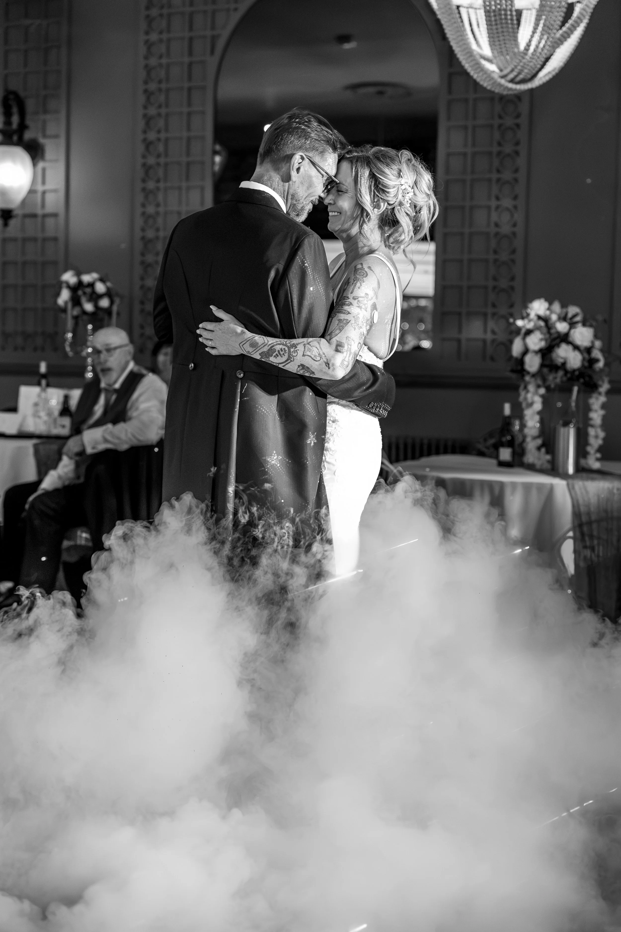 A newly married couple sharing their first dance at a wedding reception, with a fog effect on the dance floor and floral arrangements in the background.