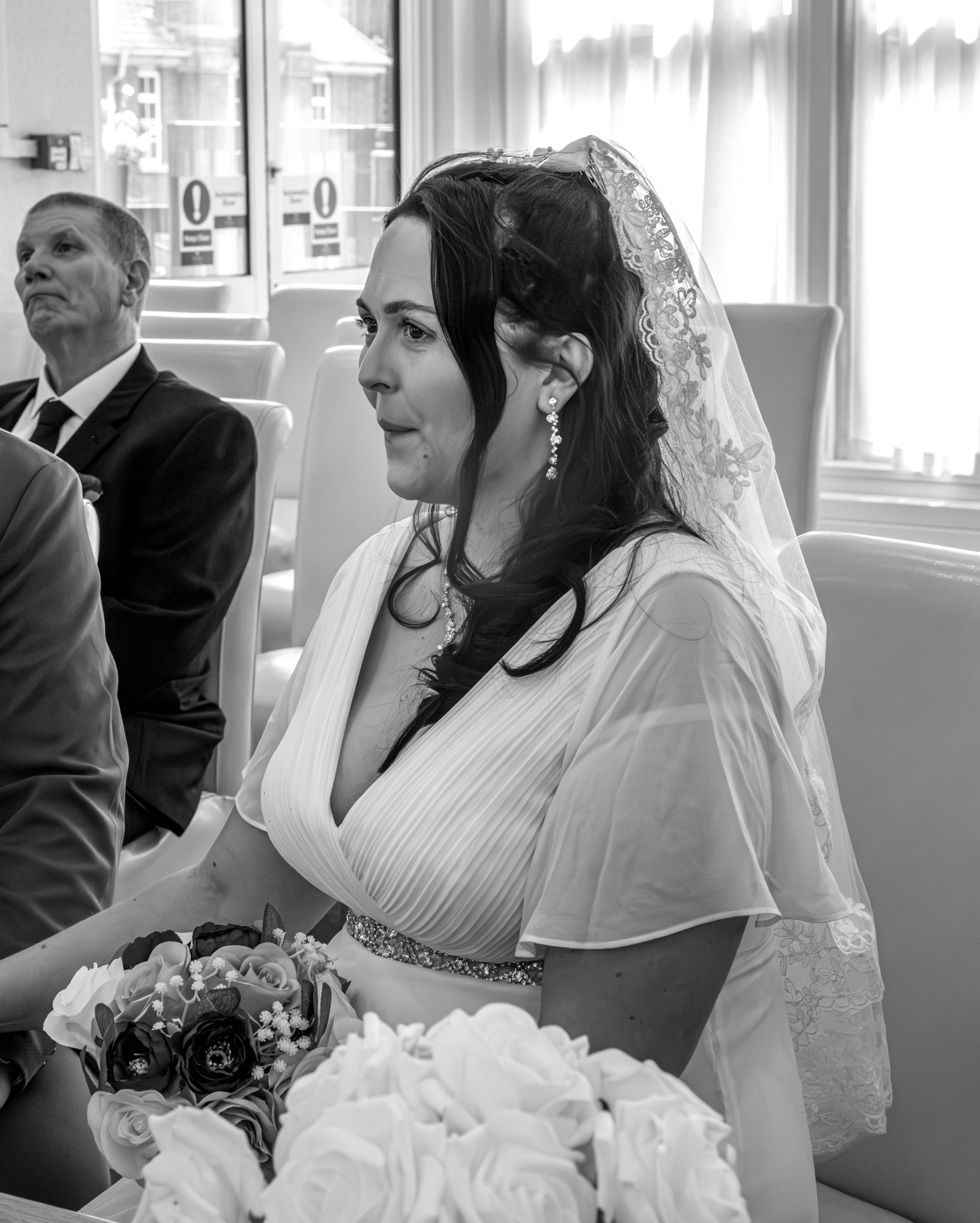 Black and white photo of a woman wearing a wedding veil and earrings, seated at a table with a bouquet of flowers, alongside a man in a suit.