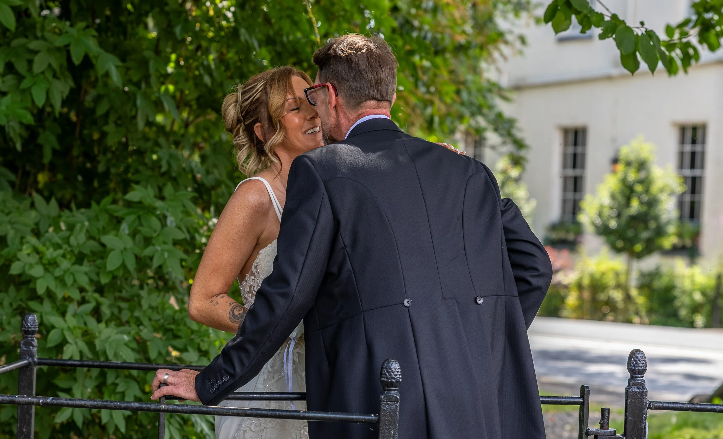 A bride and groom share a kiss outdoors on their wedding day, with greenery and a white building in the background.