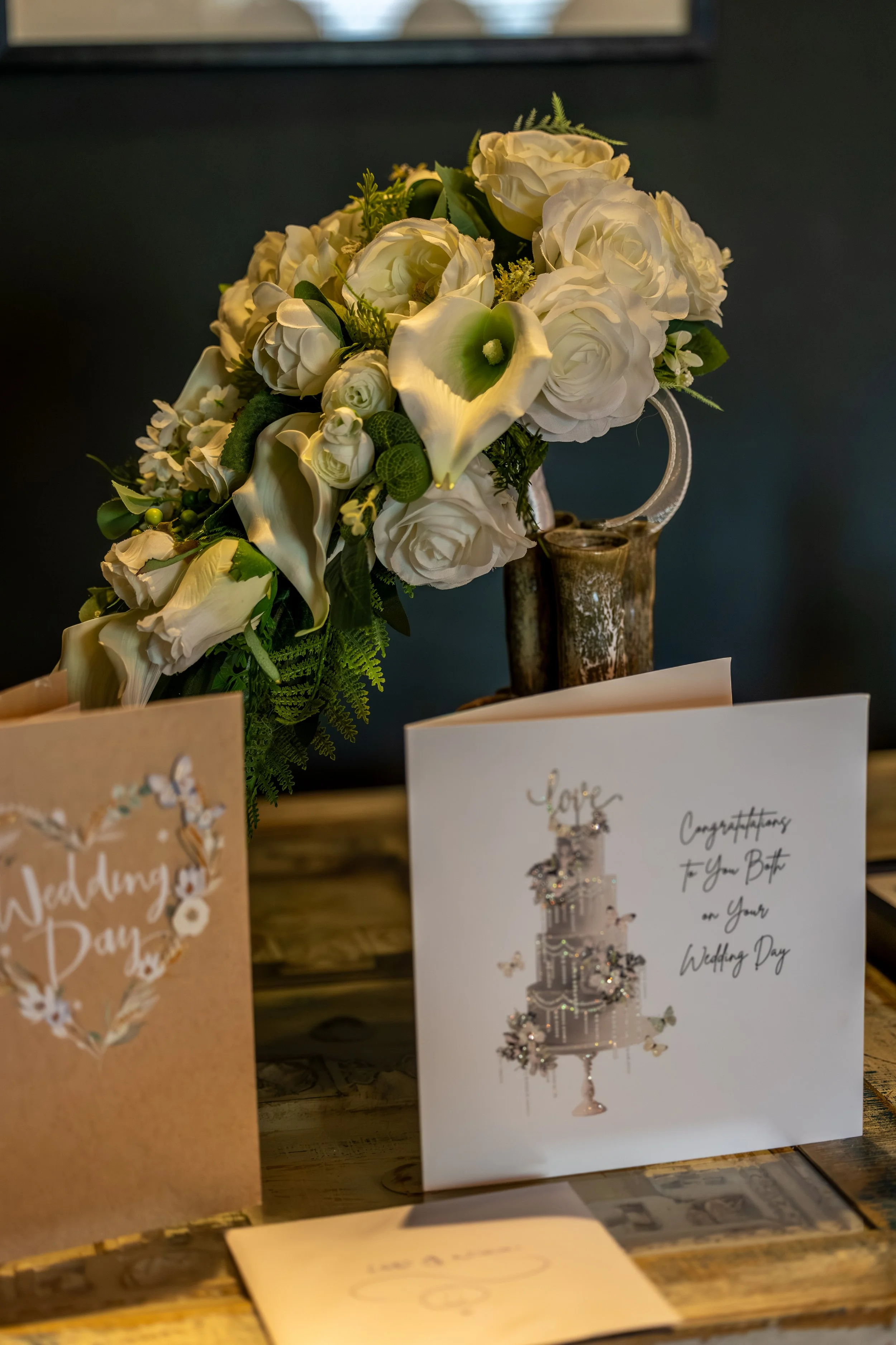 A white and green floral bouquet with calla lilies, roses, and greenery, in a rustic vase, with greeting cards and congratulatory notes nearby.