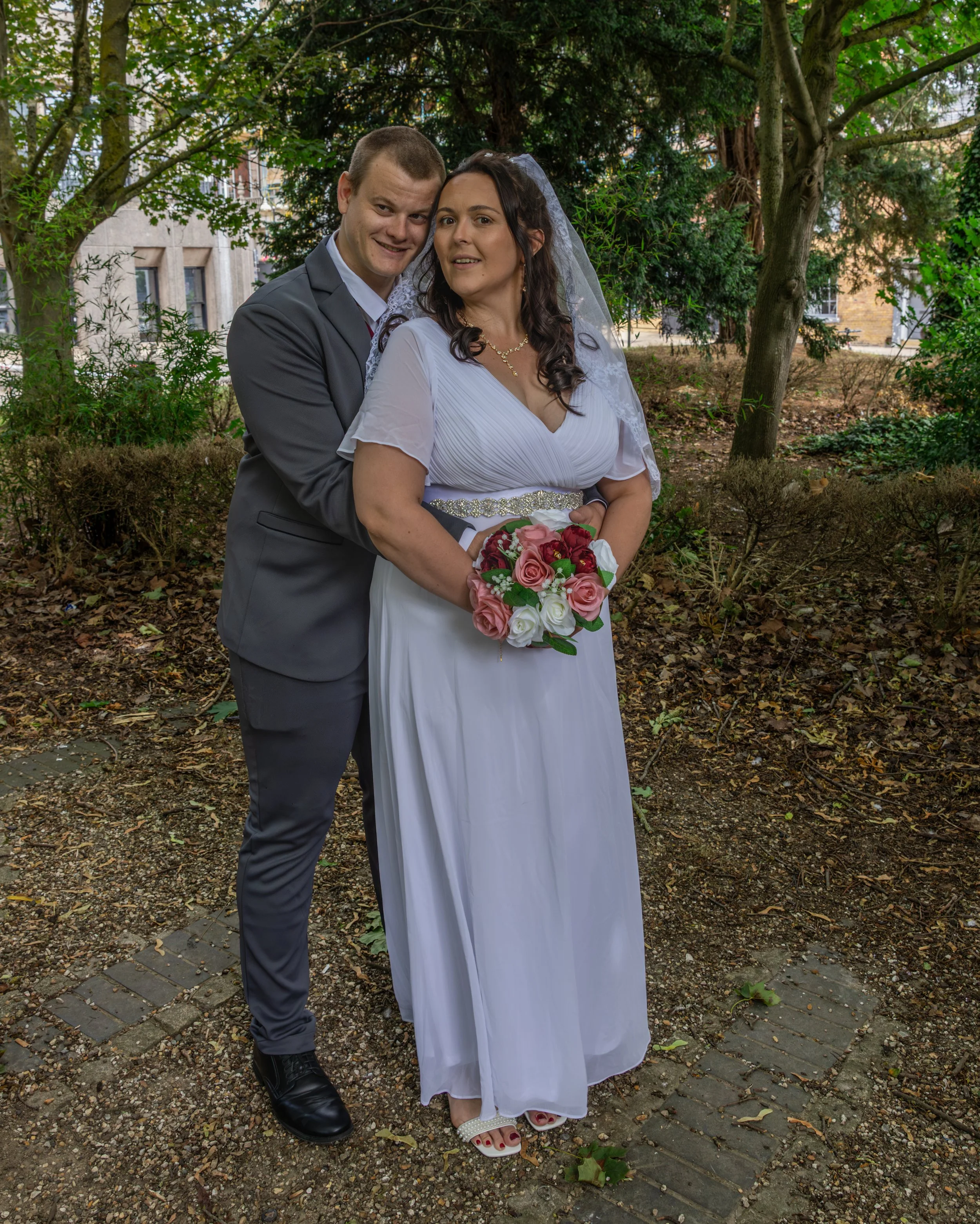 A bride and groom posing outdoors surrounded by trees and greenery. The groom is wearing a gray suit and black shoes, smiling and standing behind the bride. The bride is in a white wedding dress with a beaded belt and veil, holding a bouquet of pink 