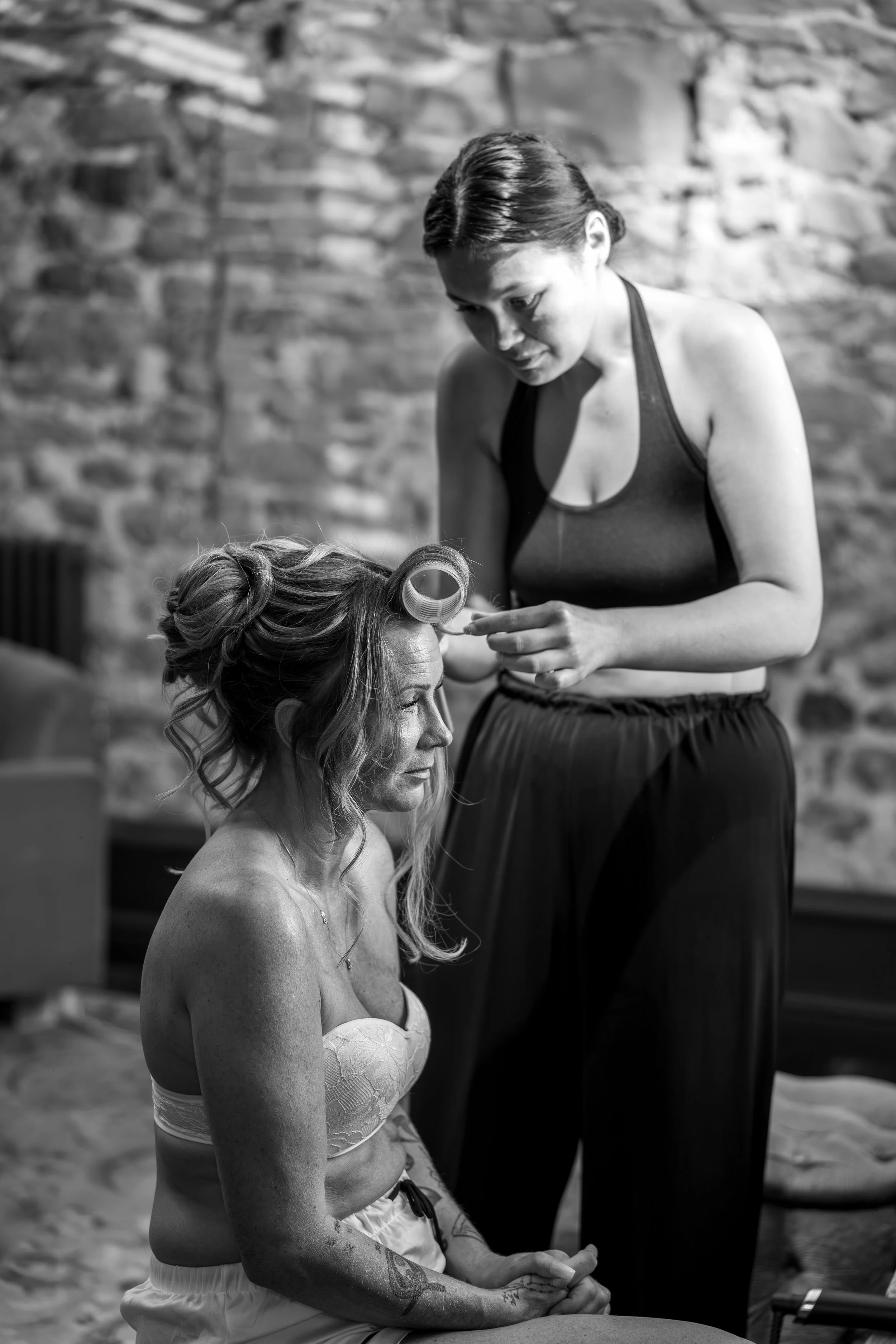 A woman with tattoos and in a strapless top is getting her makeup done by a makeup artist while she is sitting, in front of a brick wall.