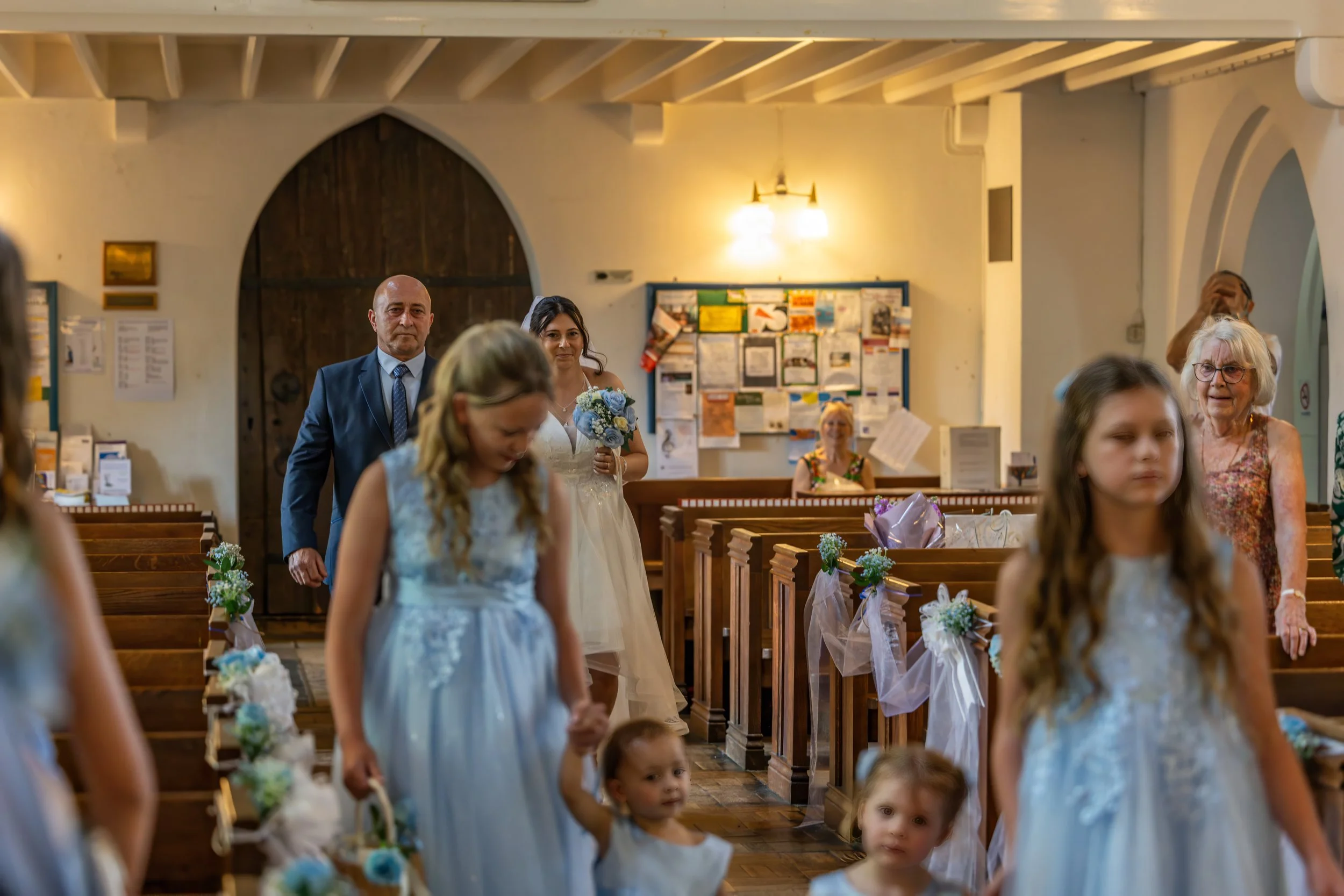 A bride in a white wedding dress and a man in a suit walking down the aisle of a church, with bridesmaids and guests visible around them.
