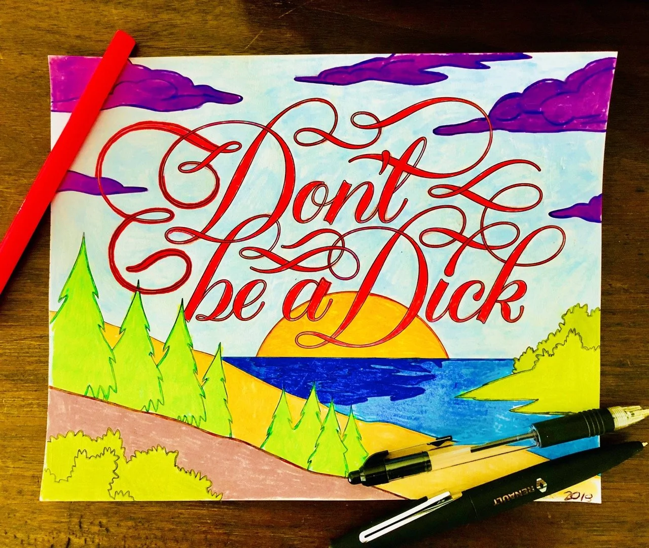  Colorful drawing with the phrase "Don't be a dick" in elegant red script, set against a landscape of trees, a lake, and a sunset. Pens and markers lie on the wooden table beside the artwork.