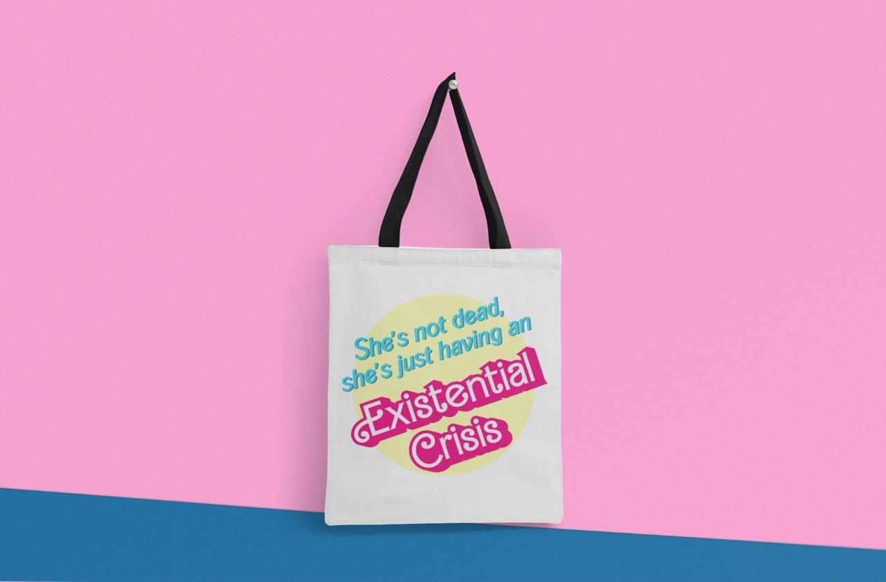 White tote bag with black handles displayed on a pink and blue background. The bag features the humorous text "She's not dead, she's just having an Existential Crisis" in blue and pink lettering with a yellow circle backdrop.