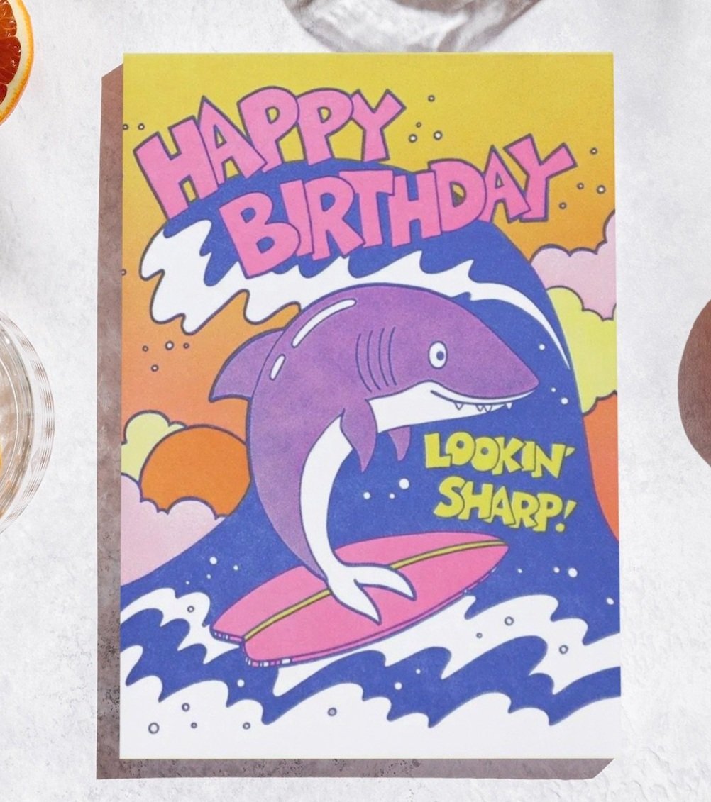 Colorful birthday card displaying a cartoon shark with 'Happy Birthday' in bold letters at the top and 'Lookin' Sharp!' in a speech bubble, set against a vibrant yellow and purple sunset background.