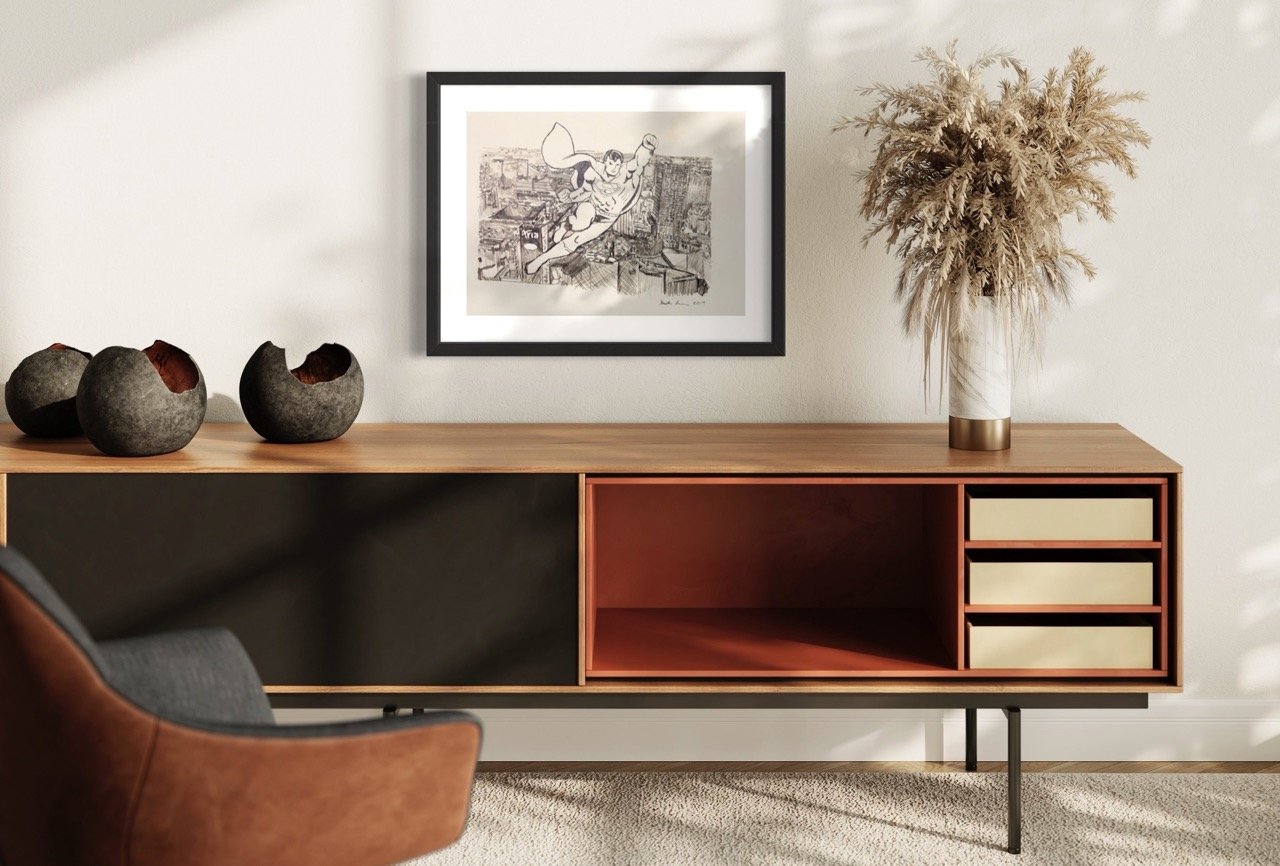 "Framed pencil drawing of a superman figure soaring over a cityscape, placed over a modern wooden credenza with decorative carved bowls and a vase of dried foliage."