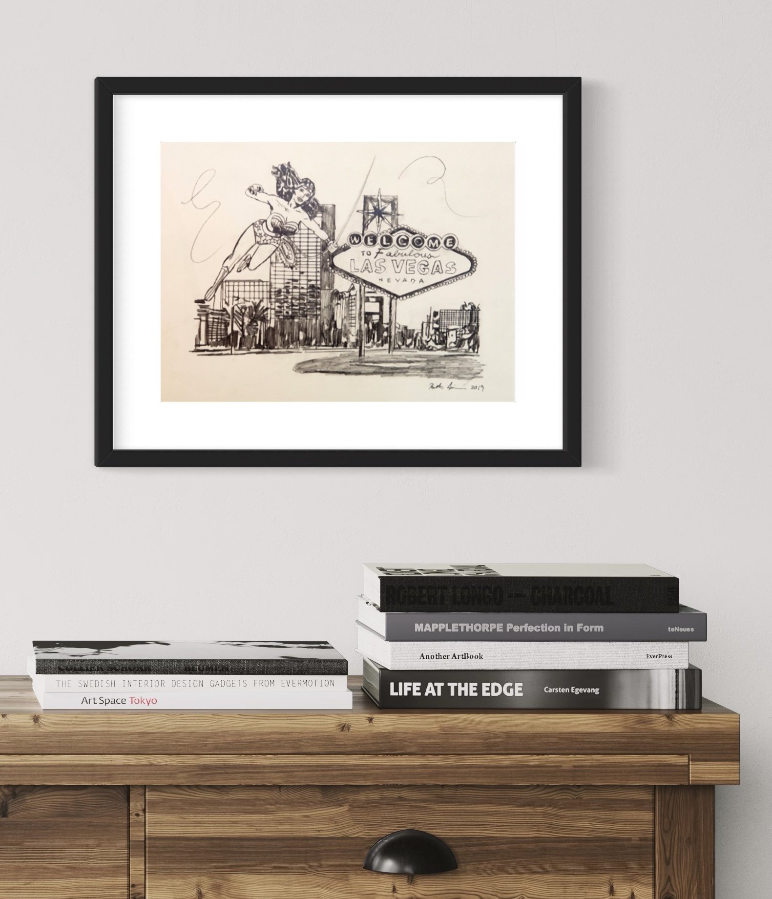 Monochrome sketch of iconic "Welcome to Las Vegas" sign with Wonder Woman, mounted in a black frame on a white wall above a wooden console