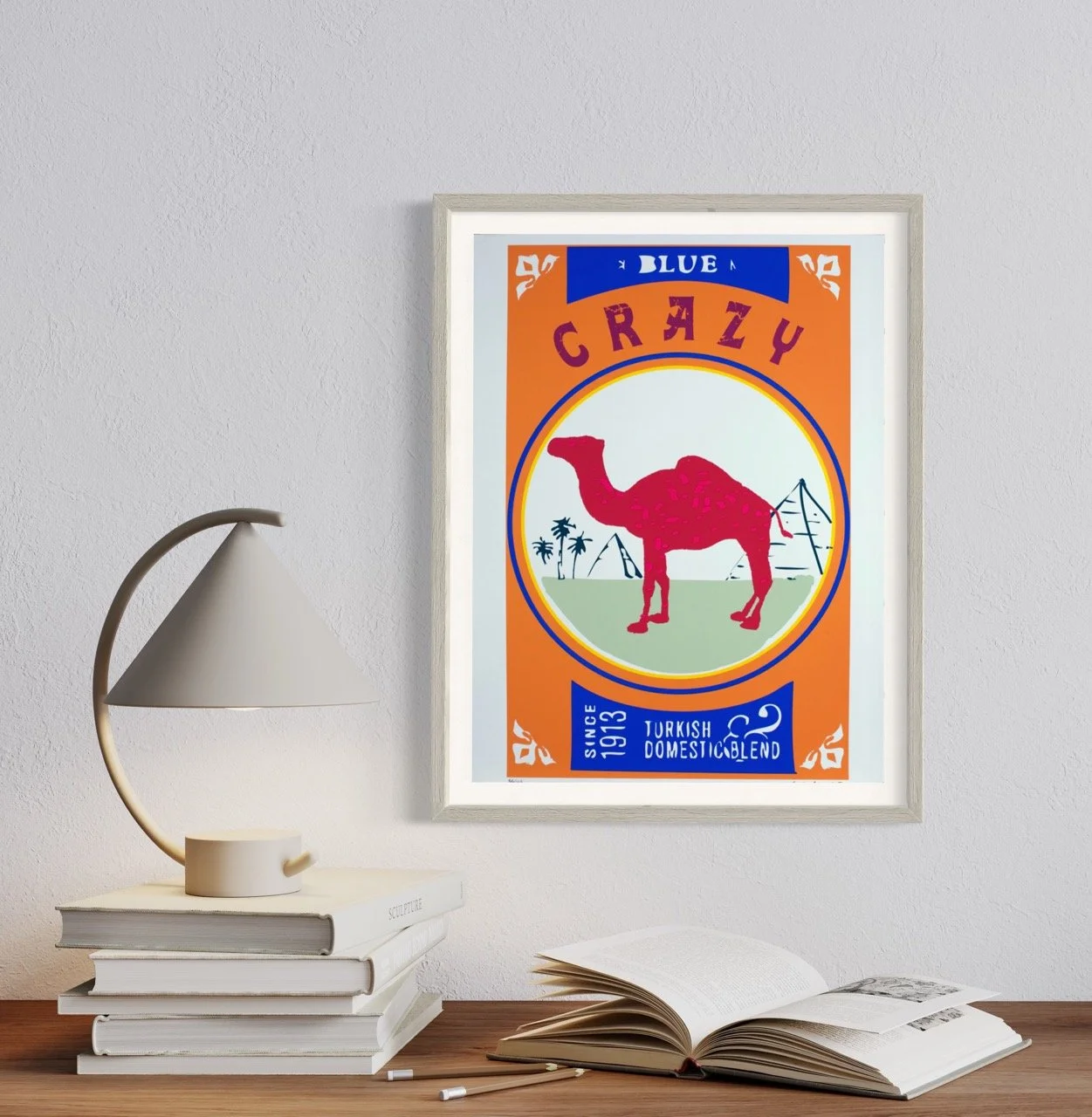 Framed pop art screenprint on a wall, depicting a pink camel with pyramids and palm trees, with text "CRAZY" and "Since 1913 Turkish Domestic Blend." The setup includes a modern lamp and a stack of books on a wooden table.
