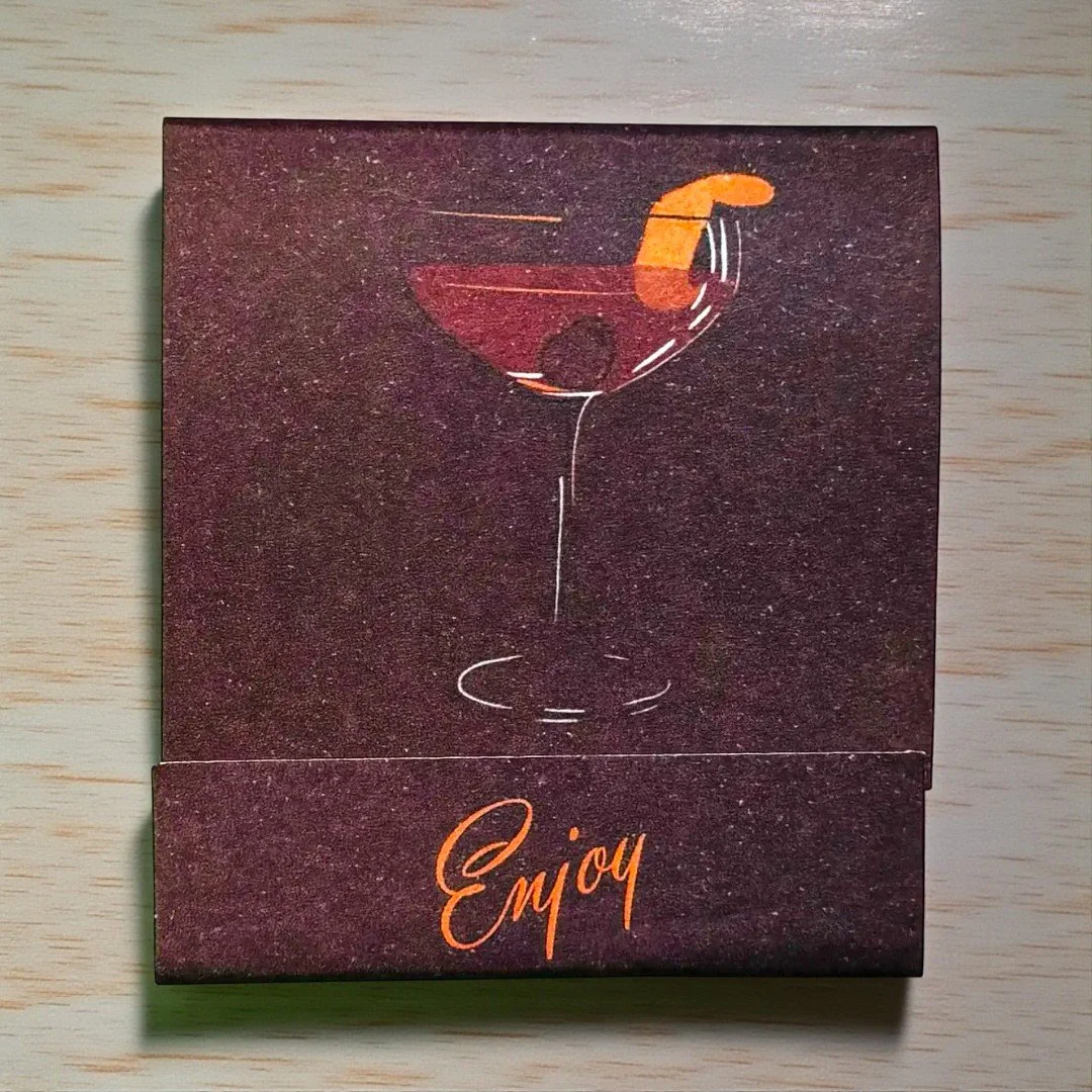 Retro matchbook featuring ingredients of a Manhattan cocktail with moody artwork and “Enjoy” text.
