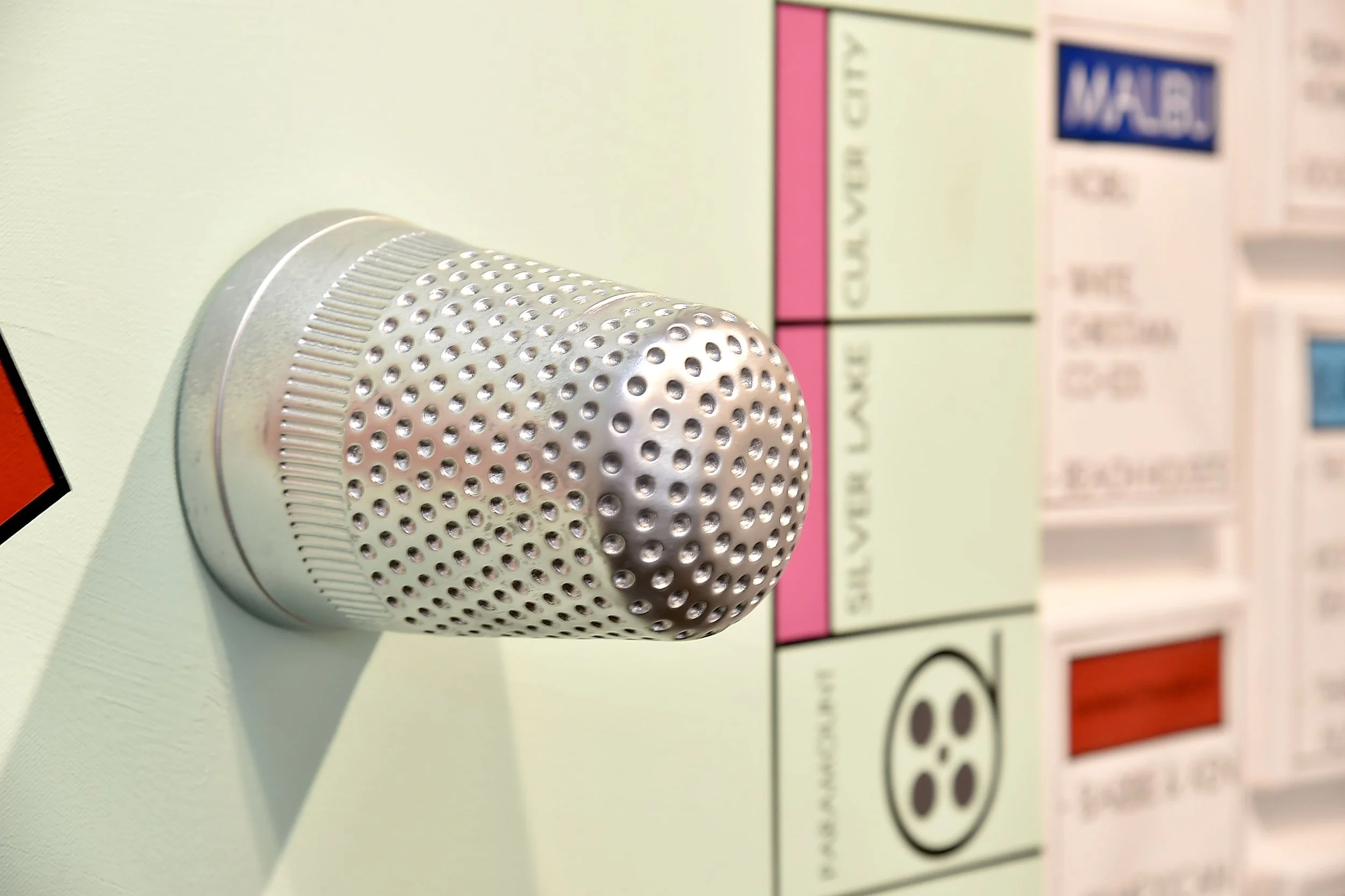 "Close-up of a large metallic thimble attached to a wall, reminiscent of a Monopoly game piece. The background features a blurred section of a game board with property names, including Silver Lake and Malibu, in pastel colors.
"