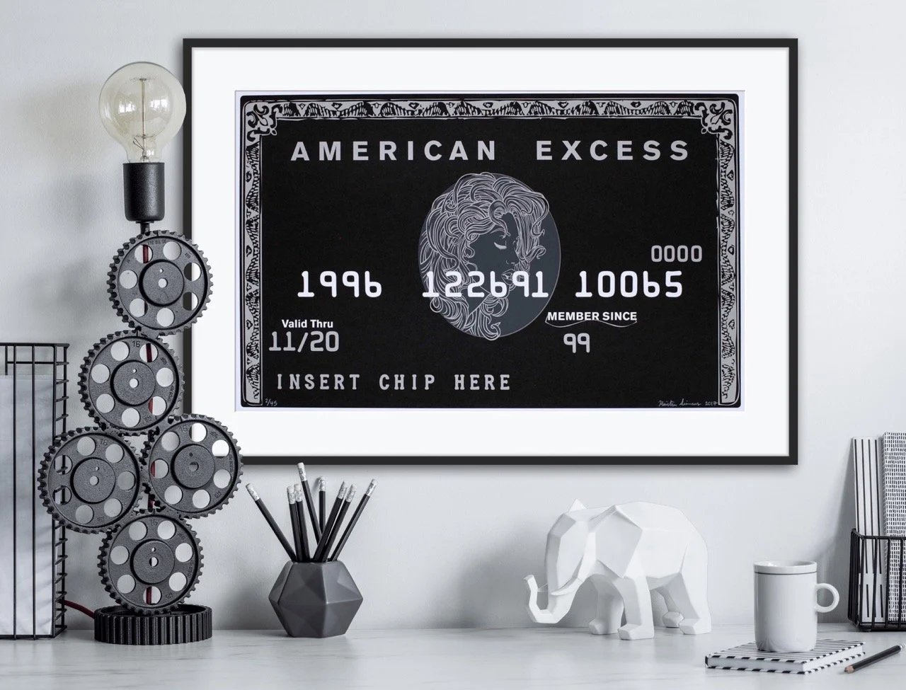 "Framed artwork titled ""American Excess"" designed like a black credit card with intricate detailing, including a female profile and the text ""Valid Thru 11/20,"" ""Member Since 99,"" and ""Insert Chip Here."" Displayed on a shelf with modern decor