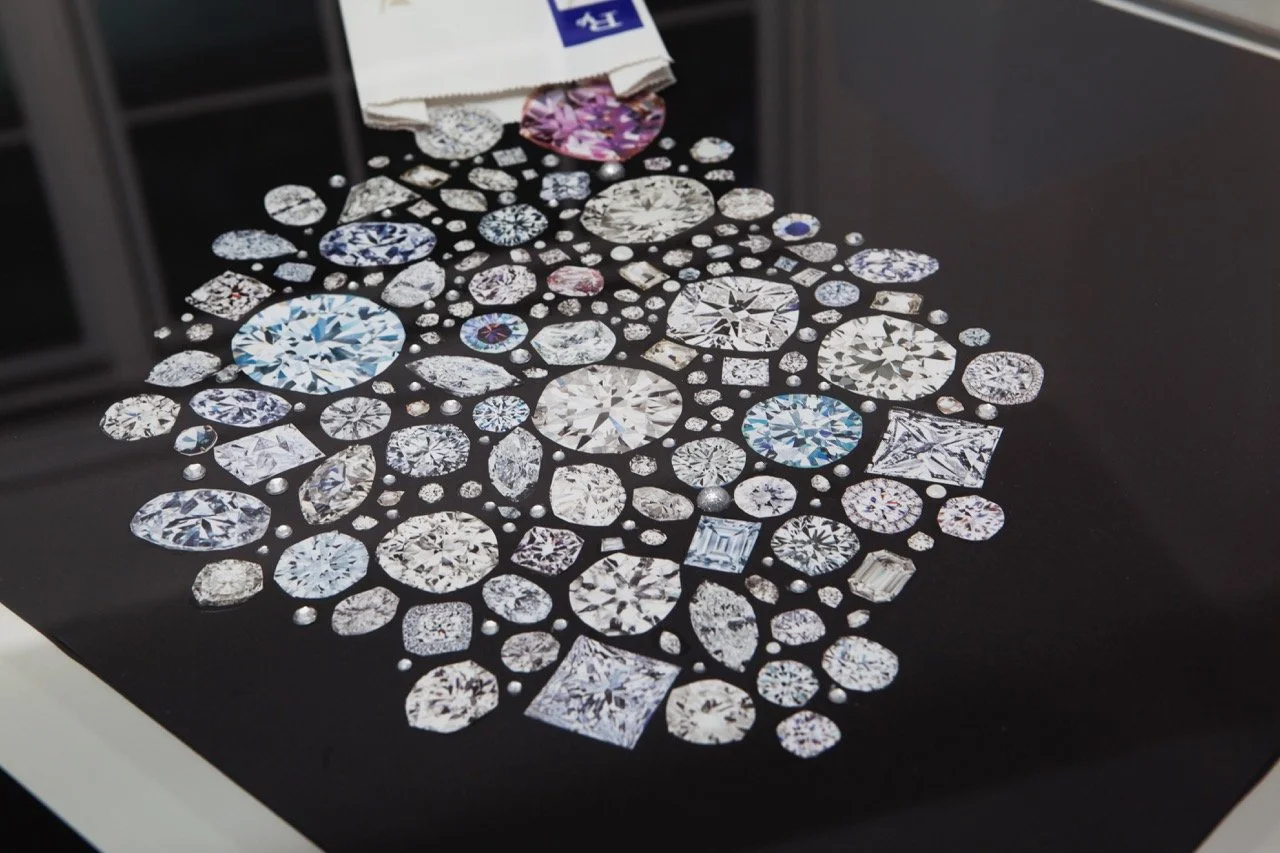 A glossy black surface adorned with a stunning array of variously cut and colored diamonds, including rounds, emeralds, and marquises. A white cloth with blue detailing lies near the top of the display.