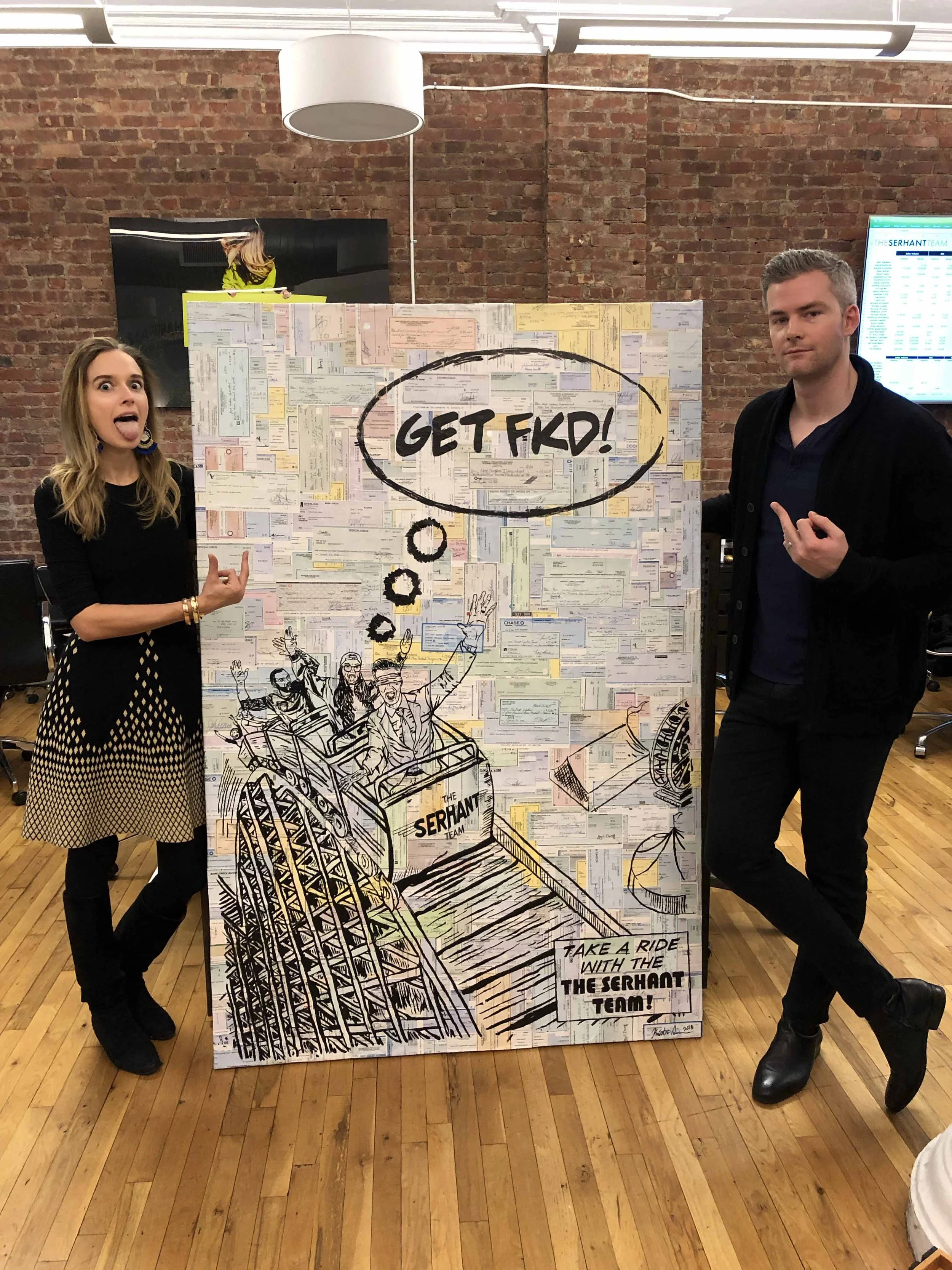 Two people stand on either side of a large artwork in an office with exposed brick walls. The artwork features a roller coaster with passengers and the phrase "GET FKD!" in a speech bubble. One person points at the art, and the other makes a gesture.