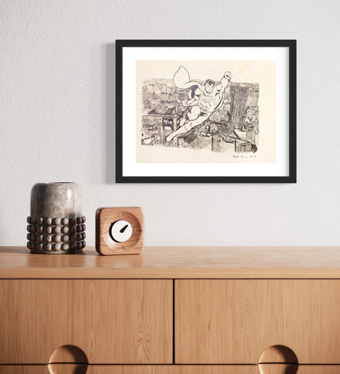 "A sketch of superman in flight over a cityscape is displayed in a frame on a light wall, above a wooden dresser adorned with a metallic vase and a wooden cube clock."