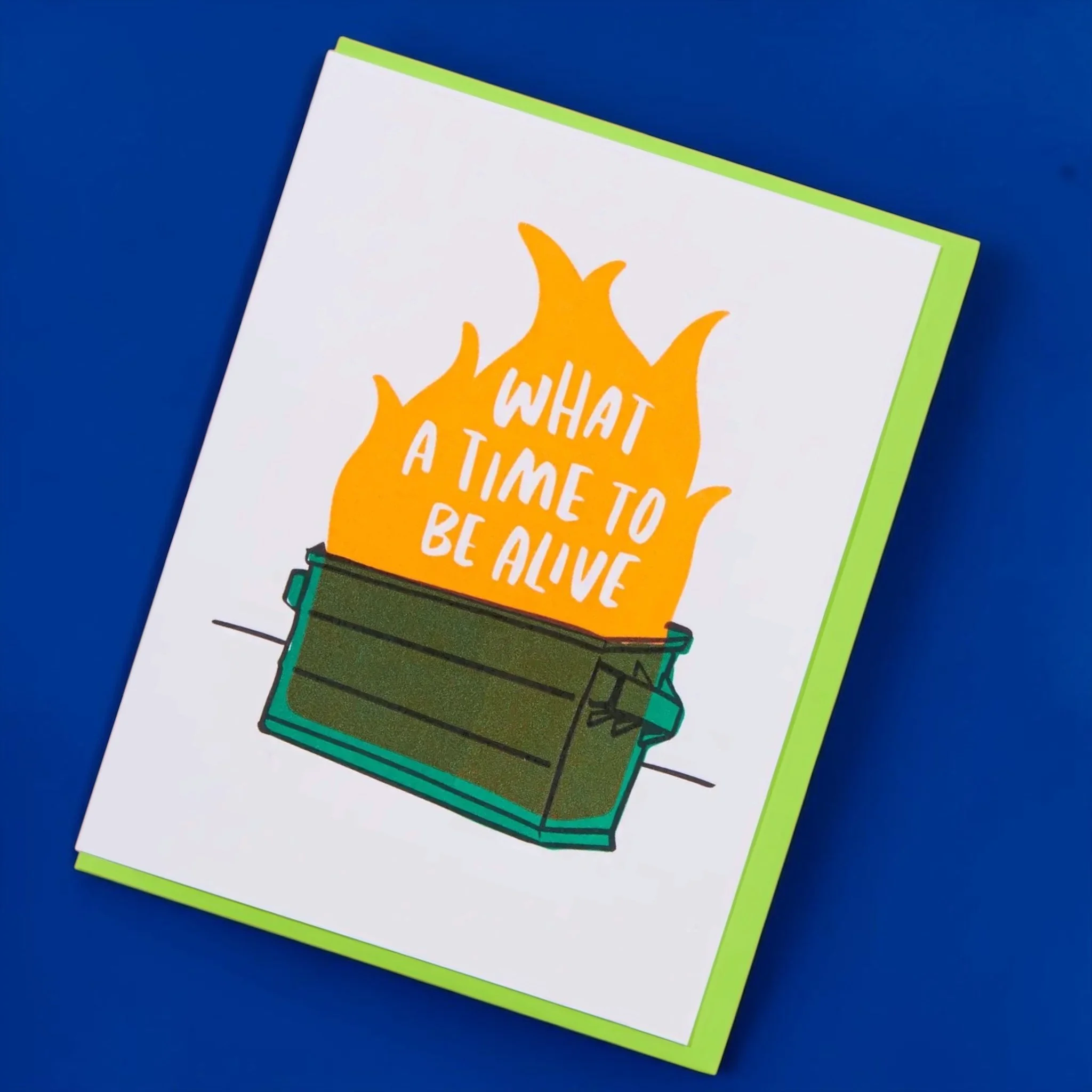 Angled view of a funny “What a time to be alive” greeting card featuring a burning dumpster illustration with a bright green envelope.