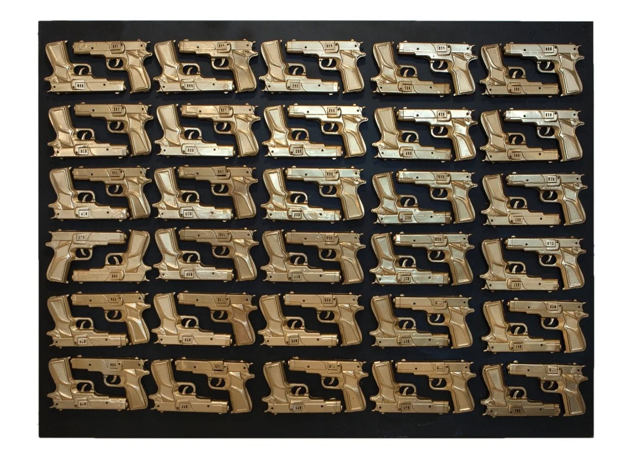 Art installation featuring rows of gold pistols arranged in a grid pattern against a black background. Each pistol is identical, creating a striking and repetitive visual effect that draws attention to the theme of the artwork.