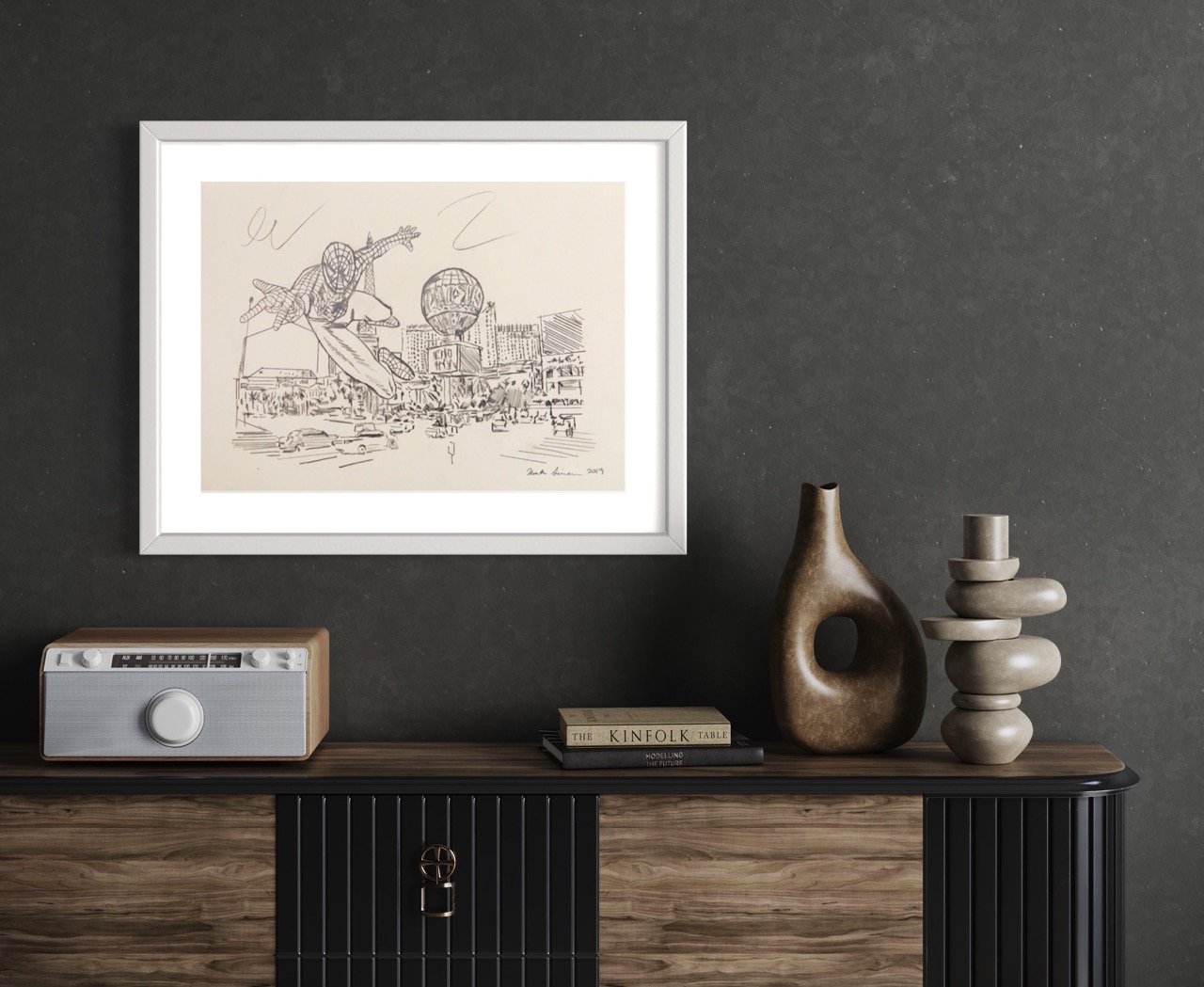 Framed ink illustration of Spiderman swinging through the Las Vegas cityscape, placed above a sideboard with a retro radio, decorative vases, and books against a dark wall.