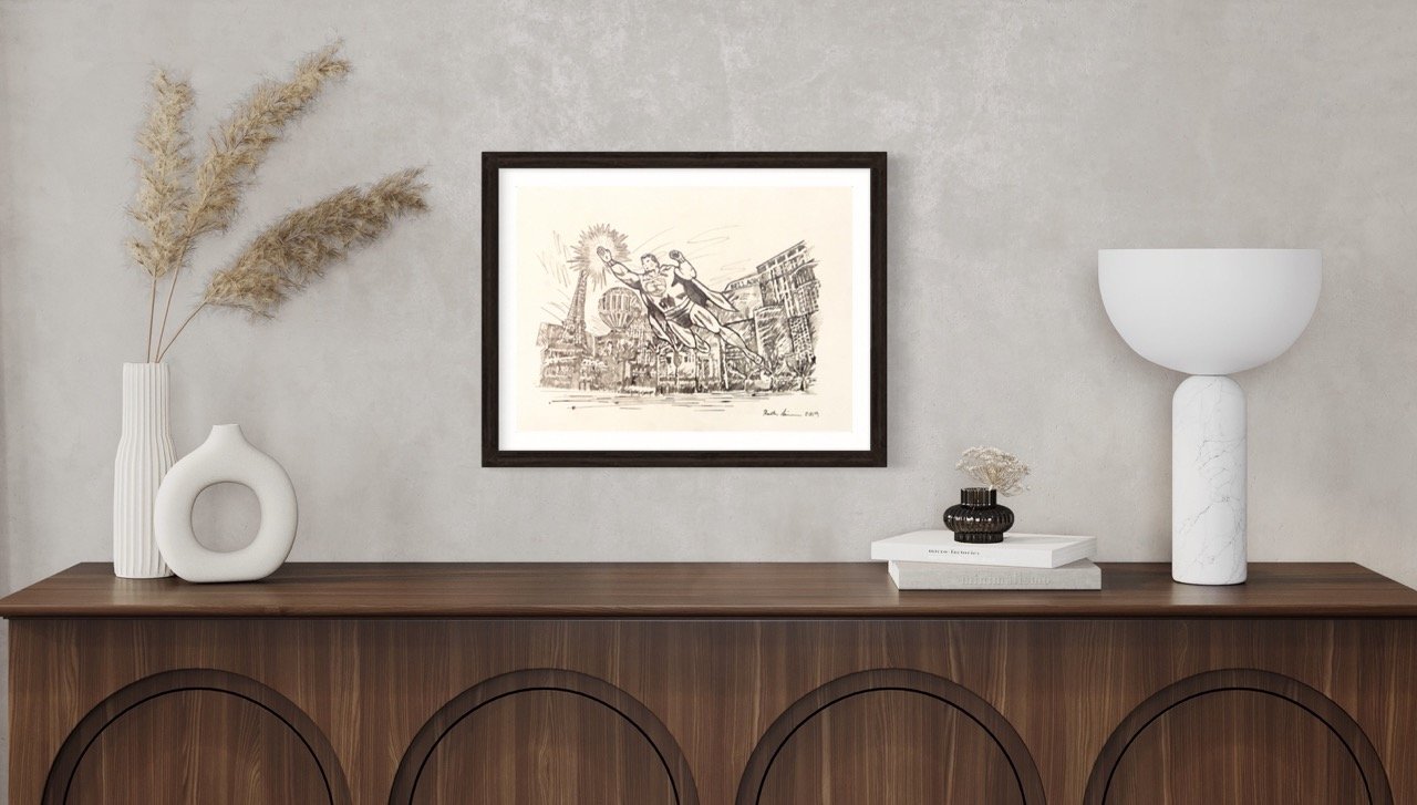 "Monochrome framed drawing superman comic with the Las Vegas cityscape, situated on a wall above a wooden cabinet adorned with a sculptural vase, pampas grass, and minimalist decor."