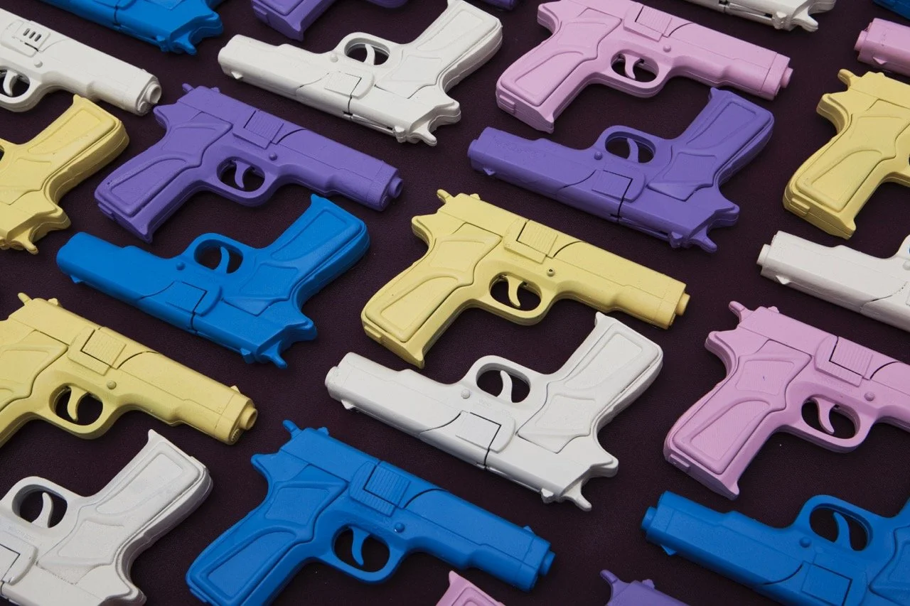 Array of toy guns in pastel colors, including blue, yellow, purple, pink, and white, laid out in a diagonal pattern on a dark background. The colorful arrangement creates a contrasting visual against the dark surface.