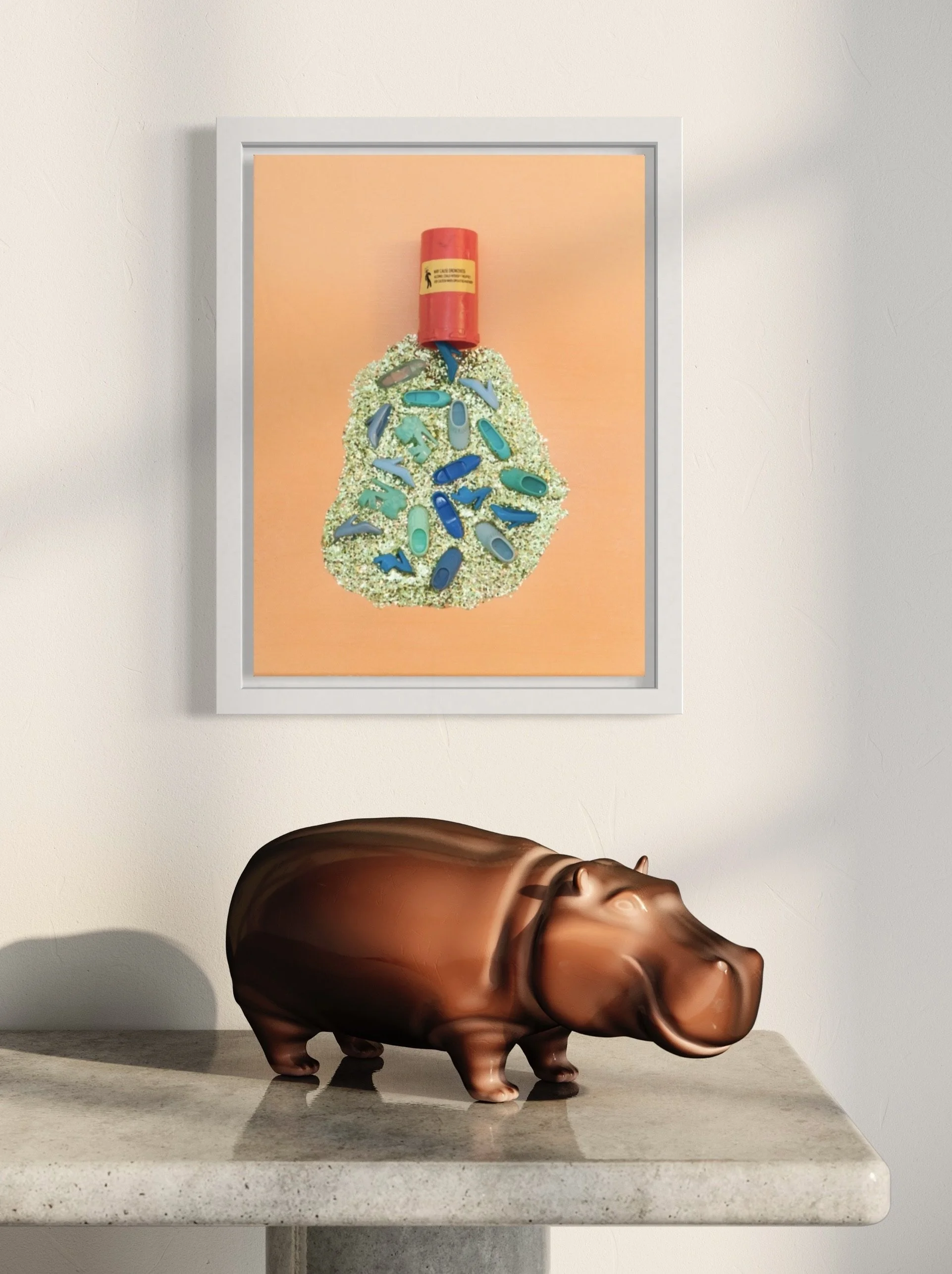 Framed art on a wall displaying a red pill bottle spilling glitter and blue and green Barbie shoes, set against an orange background. Below is a bronze hippo sculpture sits on a stone table.